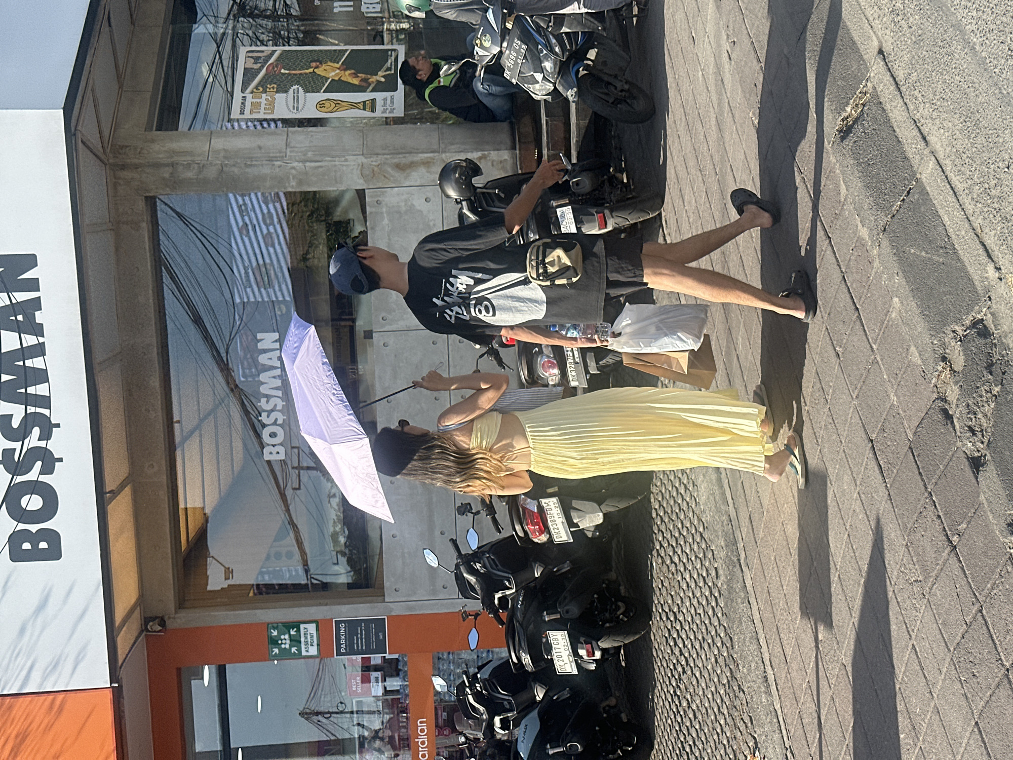 The image captures a street scene in North Kuta, Indonesia, under bright daylight. Two individuals are walking on a paved sidewalk in front of a building with a large window displaying the word "BOSS$MAN". A young woman, wearing sunglasses and a light yellow pleated dress, is holding a purple umbrella over herself and the man beside her. The man, wearing a dark t-shirt, shorts, and a baseball cap, carries a plastic bag in his left hand and a water bottle in his right. Parked motorcycles are visible lined up on the street to the left and right of the pedestrians. In the background, people are sitting on steps outside the building. The overall environment suggests a commercial area with a strong sunny ambiance.