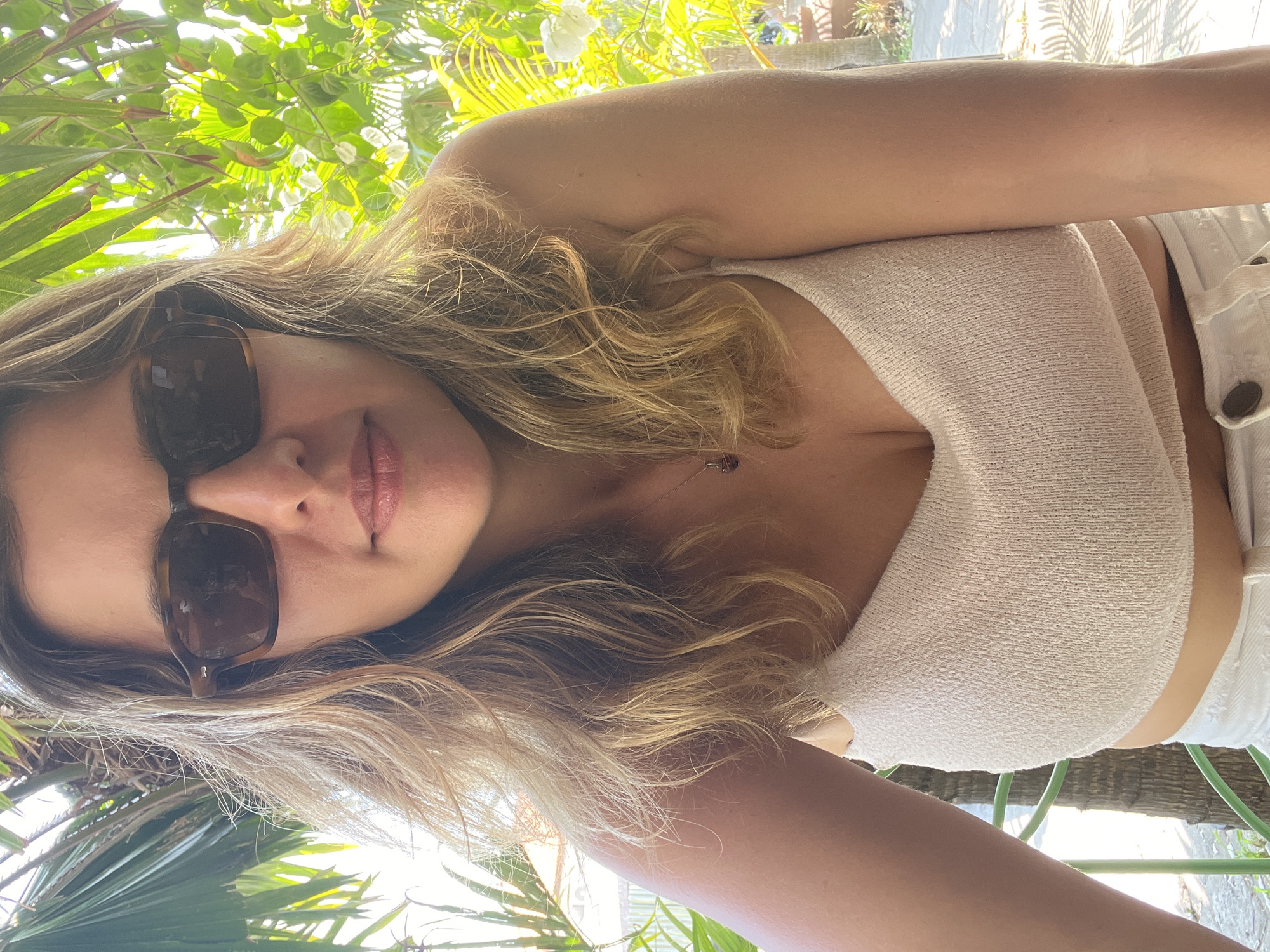 A woman with blonde, wavy hair and sunglasses is seen from the chest up. She is wearing a light-colored, textured crop top and white bottoms. Her hair is partly obscuring her face. She is outdoors, with lush green foliage and palm trees visible in the background. The lighting suggests it is daytime, with sunlight filtering through the leaves. The woman appears to be in a relaxed pose, possibly at a cafe or restaurant given the outdoor seating and vegetation. The location is specified as Mengwi, Indonesia.