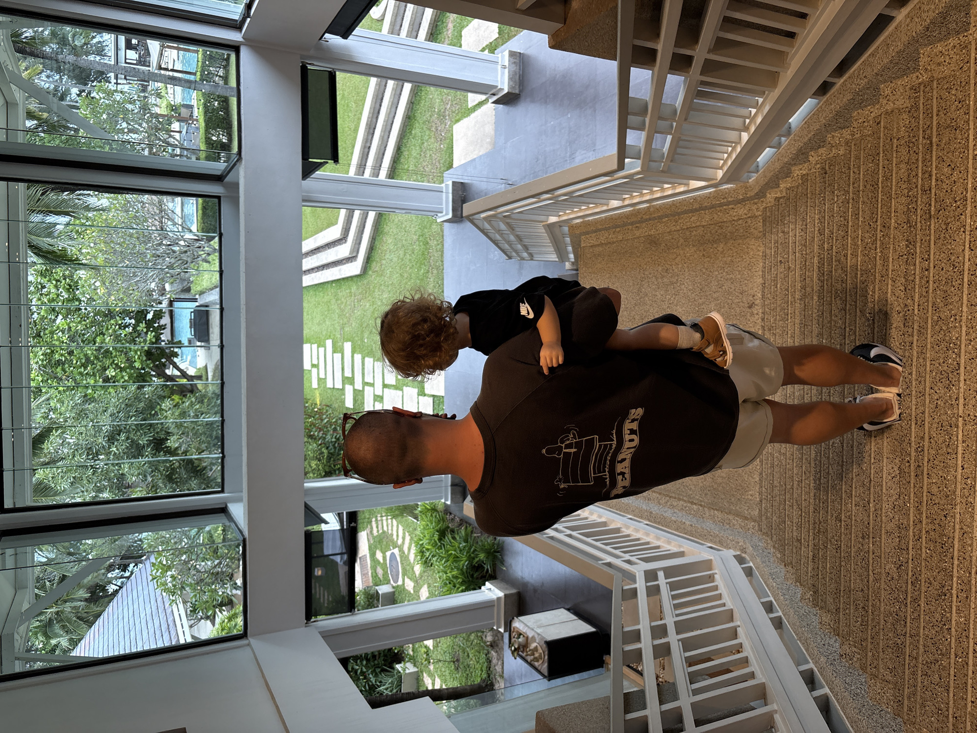 The image depicts a man carrying a young child on his shoulders inside a modern building with large windows overlooking a lush green outdoor space. The man is wearing a dark t-shirt with a graphic print of Snoopy and the word "PEANUTS" visible, and light-colored shorts. The child, with curly blonde hair, is wearing a black t-shirt and brown sandals. They are standing on a textured floor, possibly terrazzo or concrete, with a white staircase to their right. Through the large windows, a swimming pool, palm trees, and manicured gardens are visible, suggesting a tropical location. The scene appears to be indoors, looking out. The lighting suggests it could be daytime. The overall mood is peaceful and familial.