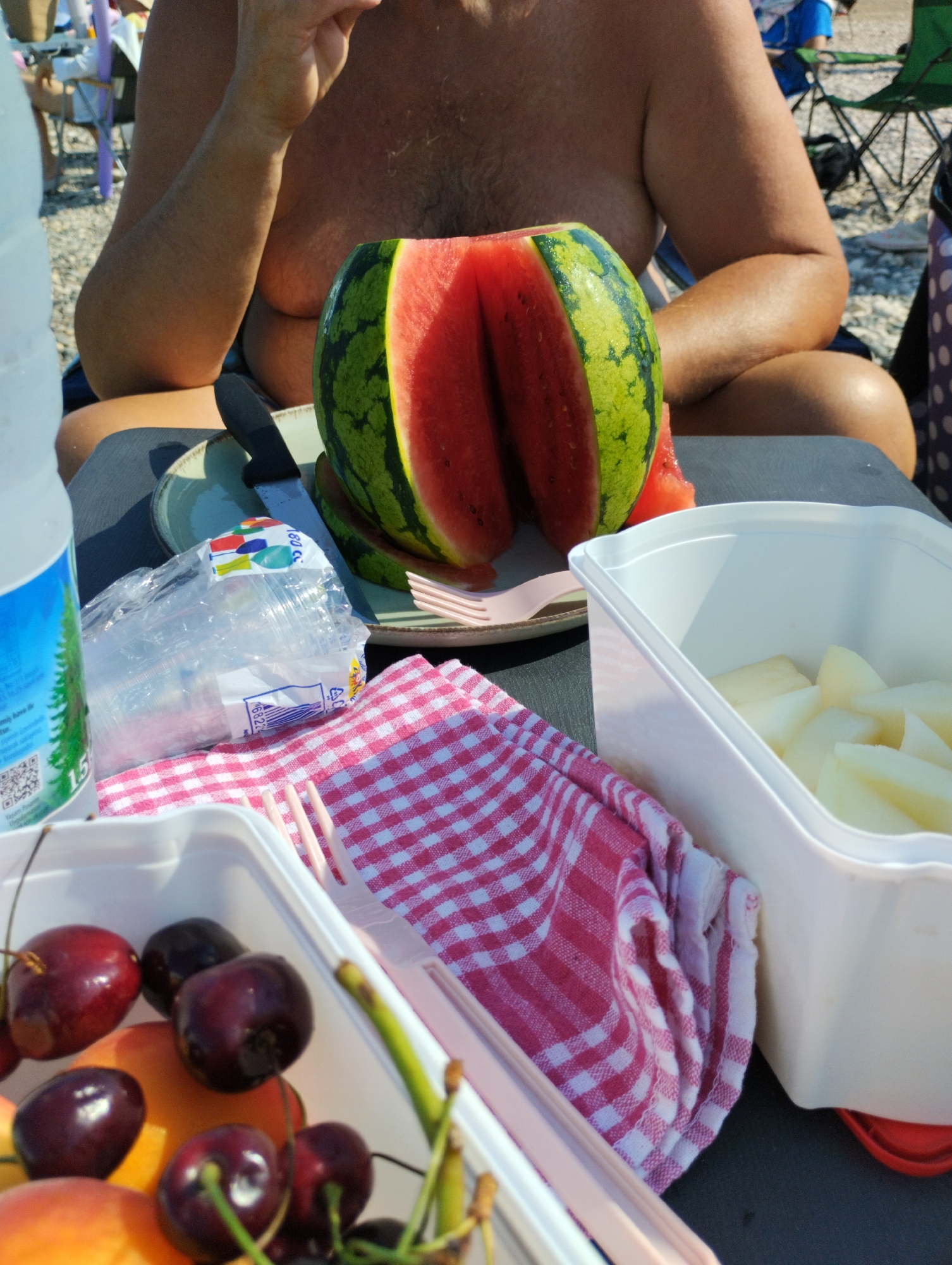 The image depicts a close-up view of a picnic spread on a table, likely at a beach or outdoor recreational area, possibly in Erdemli, Turkey given the context of a picnic. In the foreground, a white plastic container holds sliced fruit, appearing to be melon, and another container holds cherries and sliced peaches. A red and white checkered cloth is draped nearby. Behind these, a partially cut watermelon with vibrant red flesh and green rind is visible, resting on a plate with a knife. To the left, a plastic bottle of water is present, and a package of disposable cups is also in view. A person's torso and arms are partially visible in the background, suggesting they are sitting and possibly eating. The environment appears to be sunny, indicated by the lighting and the relaxed outdoor setting with hints of beach chairs and sand in the distance. The overall scene conveys a casual, summery picnic atmosphere.