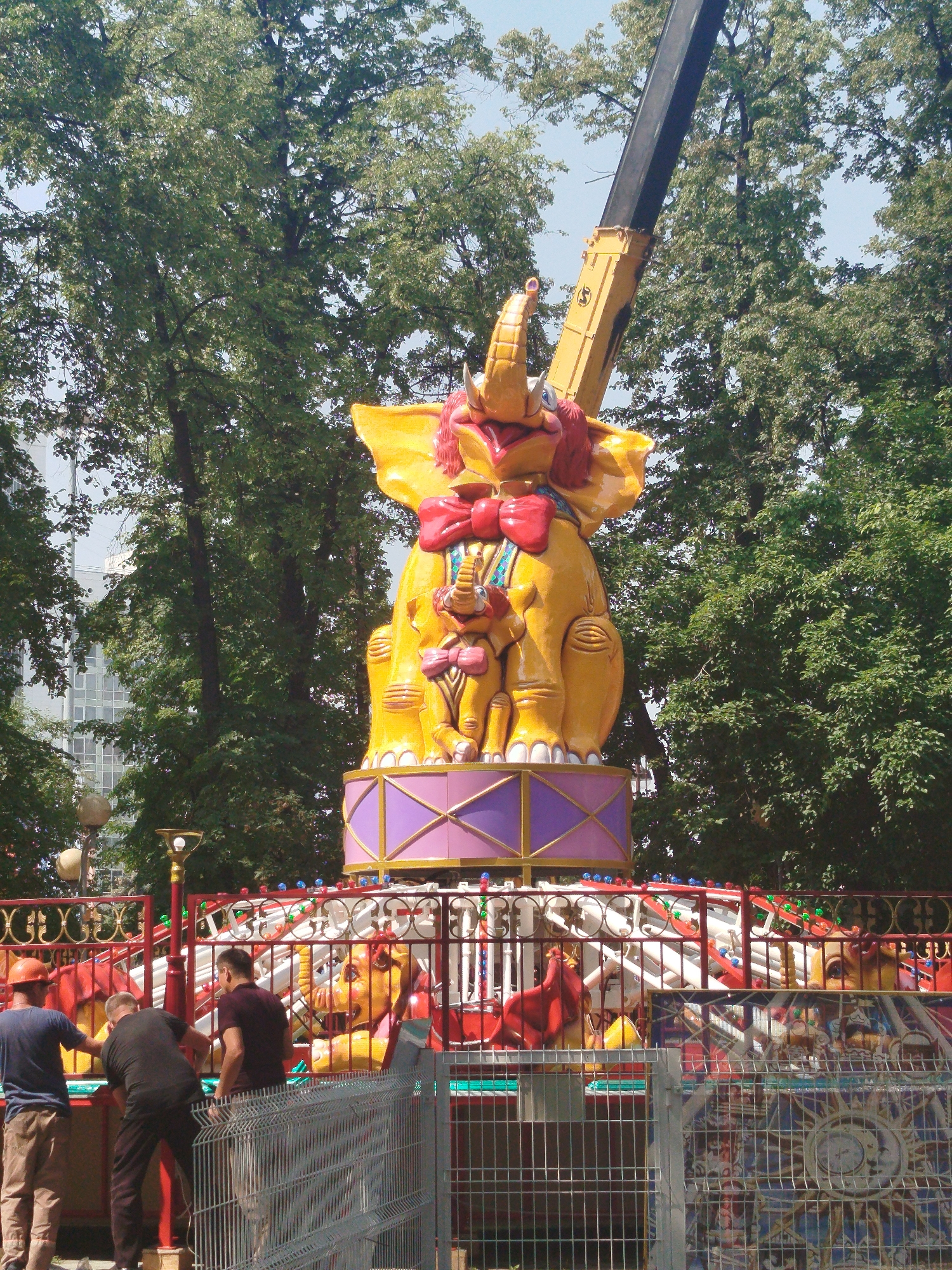 In Tyumen, Russia, a bright yellow amusement ride shaped like a large elephant with a red bow is the central focus. The elephant is positioned atop a purple and gold platform. Below the elephant, the amusement ride has a colorful metal fence with red, white, and blue lights. In front of the fence, three men are visible. One man on the left is wearing a blue t-shirt and beige pants, with an orange hard hat. The man in the middle is wearing a black t-shirt and black pants. The man on the right is wearing a dark red t-shirt and dark pants. They appear to be working on or inspecting the ride. Behind the amusement ride, lush green trees and a glimpse of a modern building are visible against a clear blue sky.