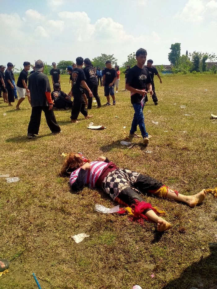 The image depicts an outdoor scene in a dry grassy field within the city of Celep, Indonesia, under a partially cloudy blue sky. In the foreground, a person is lying supine on the ground with eyes closed, dressed in traditional Indonesian attire consisting of a red and white striped long-sleeved shirt, black and white checkered shorts over dark trousers, and red and yellow fabric ornamentation around their shins and ankles. A brown, shaggy headpiece or wig covers their head and face. The person's feet are bare and appear dirty.
Scattered across the dry grass in the foreground and midground are various pieces of litter, including plastic bags, paper plates, and plastic cups. A blue stick-like object is visible in the bottom left corner.
In the midground, a group of approximately 15-20 males, predominantly young adults, are standing and walking across the field. Most are wearing black t-shirts, some with graphic designs, and dark trousers. One individual in blue jeans and a black t-shirt with a patterned sash is walking towards the right, looking towards the viewer. Another individual wearing a black t-shirt with orange visible on the sleeves is walking away from the camera. The group appears to be observing or participating in an event.
The background shows a line of green trees and sparse vegetation, with a light-colored building partially visible through the trees on the right side. The ground throughout the scene is characterized by dry, yellowish-green grass with patches of exposed dirt.