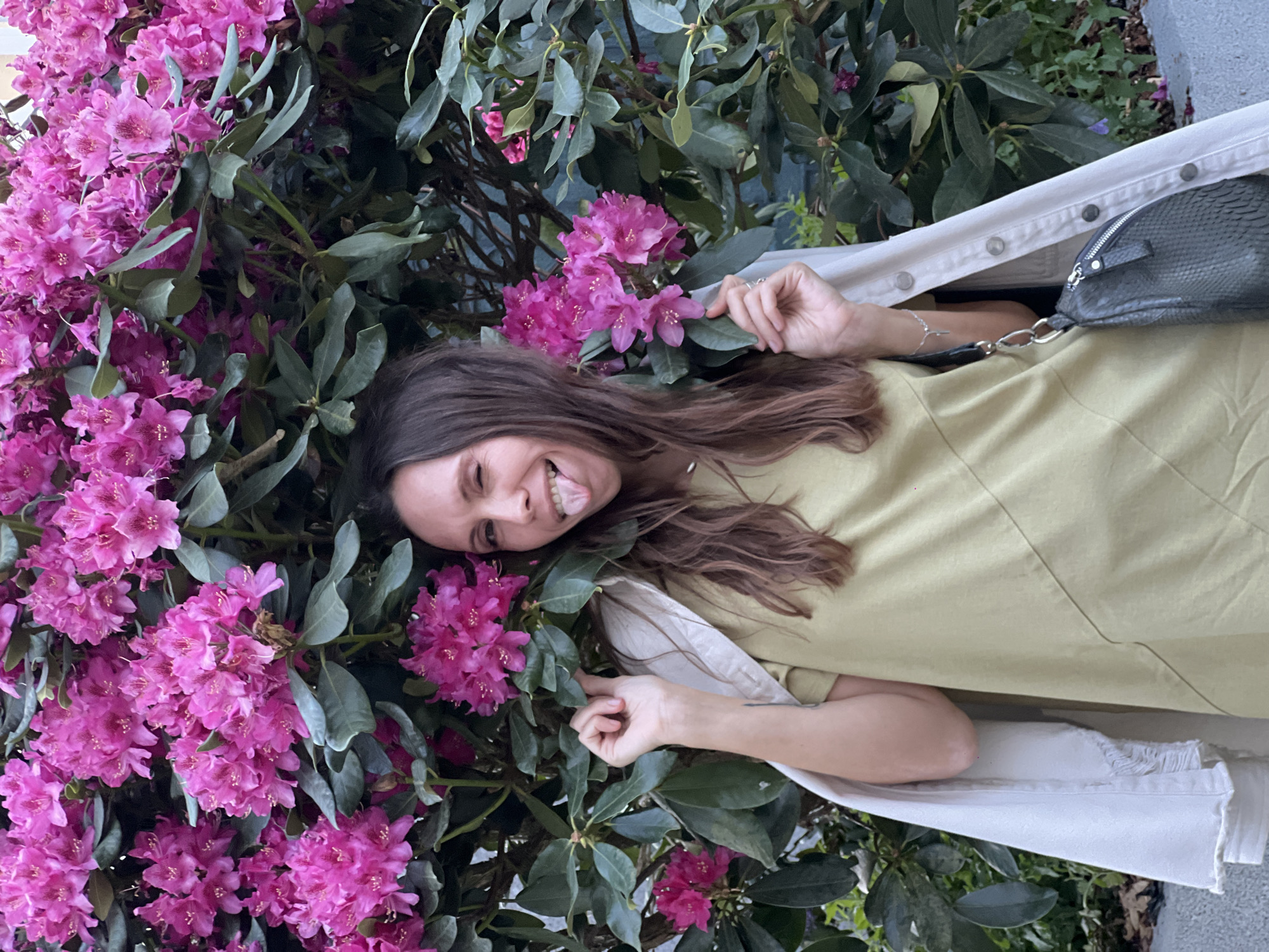 A young adult female is depicted standing in front of a dense flowering bush of vibrant fuchsia rhododendrons with dark green foliage. She is performing a deliberate wink with her right eye and extending her tongue outwards to her right. Her left hand is observed lightly touching a cluster of the pink flowers. A light beige or off-white denim jacket is draped over her shoulders, with her right hand partially holding it. She is wearing an olive-green short-sleeved dress. A silver-toned bracelet with a small cross charm is visible on her left wrist. A dark grey, textured handbag with a silver chain strap hangs from her right side. Her long, dark brown hair is styled straight and falls over her shoulders. The background behind the flowering bush suggests a dark blue or green wall or fence. This scene is situated in Moscow, Russia, likely within a cultivated green space such as a park or botanical garden.