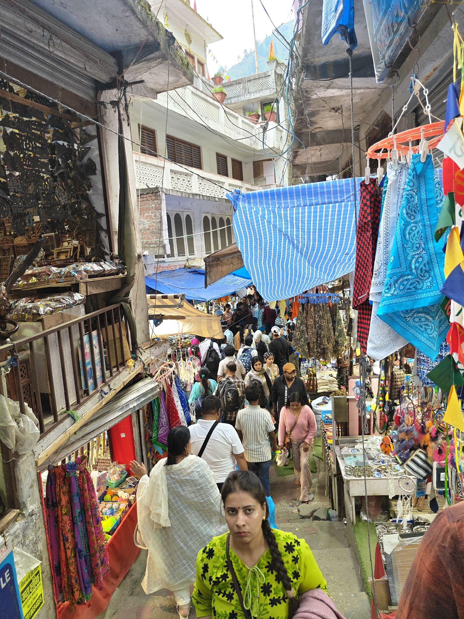 This image captures a vibrant and bustling market scene within a narrow alleyway, likely in Kasol, India, given the mountain context and general atmosphere. The lane is crowded with people, mostly adults, navigating their way through the throng, suggesting a lively shopping or pilgrimage route.
On both sides of the alley, small shops and stalls are packed with various goods. On the left, a shop displays numerous metal crafts or decorative items, possibly religious or souvenir-related, some packaged in plastic. Further down on the left, colorful scarves are hung for sale. The right side of the alley features brightly colored bandanas, some blue with white patterns, and checkered red and black ones, hanging from clotheslines. Dreamcatchers and other craft items are also visible on display tables.
Overhead, blue and white striped fabric and solid blue tarpaulins are strung across the alley, providing shade from the sun. Colorful triangular prayer flags, typical of Himalayan regions, are also strung, adding to the festive atmosphere. The buildings framing the alley are multi-storied with white and pastel facades, some featuring decorative balconies and arched windows, hinting at traditional architecture. Tangled electrical wires are visible overhead, a common sight in such densely populated areas.
In the foreground, a woman in a bright lime-green top with black floral patterns and braided hair looks directly at the camera with a neutral, perhaps slightly wary, expression. Behind her, another woman in a white outfit walks away. The ground appears to be an uneven mix of steps and paved surfaces, contributing to the dynamic feel of the scene. A mountain can be faintly seen in the background through an opening between buildings.
Visible text in the image includes fragments like "LE", "ock", "जाता कचीनक", "जीउन भर के लिए असम", a phone number "7018034055", and "JIVE MINDS #AREN".