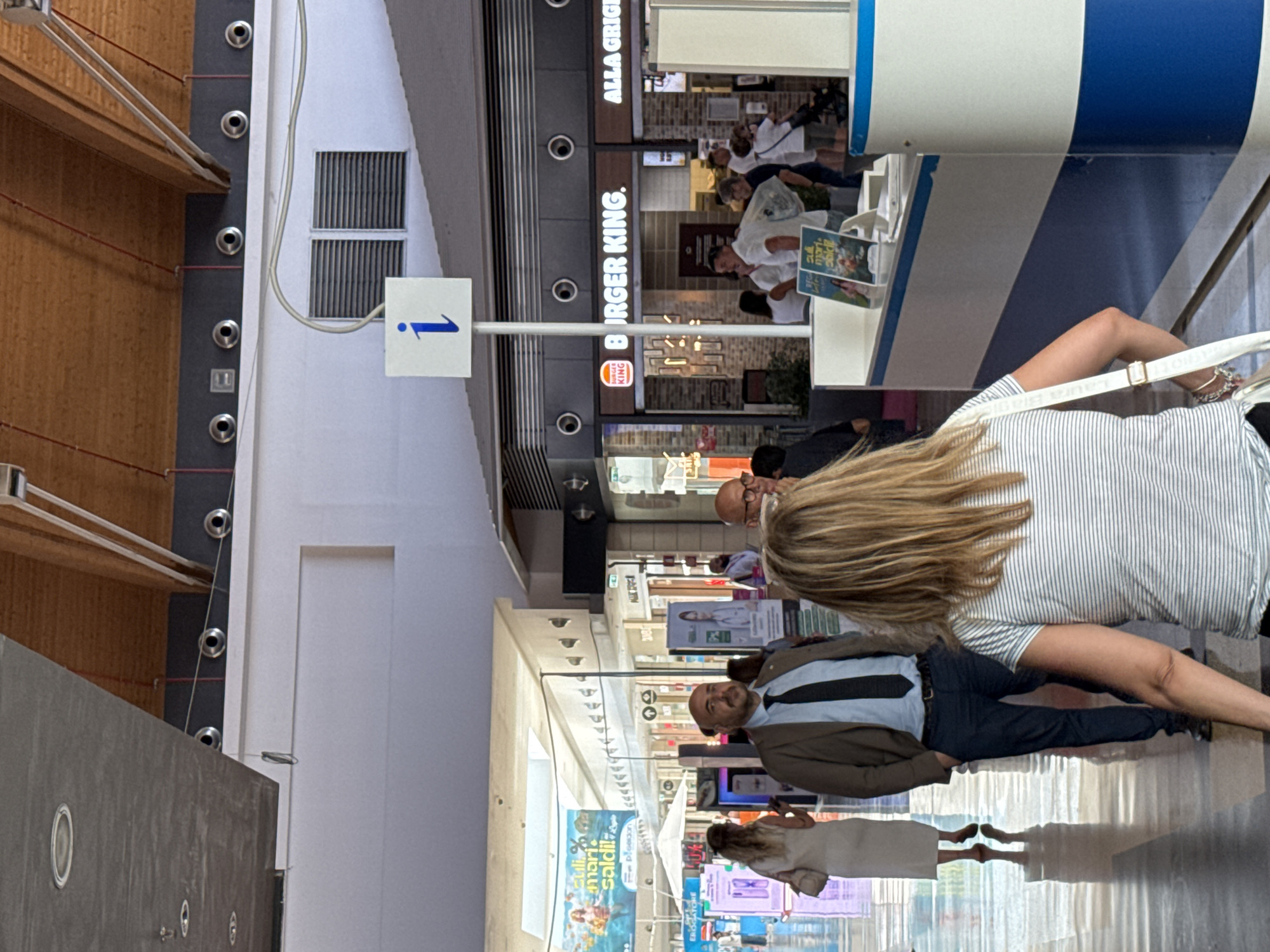 This image depicts an indoor shopping mall environment, specifically within the Auchan shopping center in Carini, Italy. The scene captures a public walkway with various shops and people. In the foreground, a woman with blonde hair, wearing a striped t-shirt and a white crossbody bag, is seen from behind, facing away from the camera. To her left, a man in a suit and tie stands with his hands in his pockets, looking forward. Further back, a Burger King restaurant is visible with patrons queuing and standing nearby. Other shops and advertisements line the walkway, and the architectural features of the mall, including a wooden ceiling and lighting fixtures, are apparent. The overall impression is of a bustling commercial space.