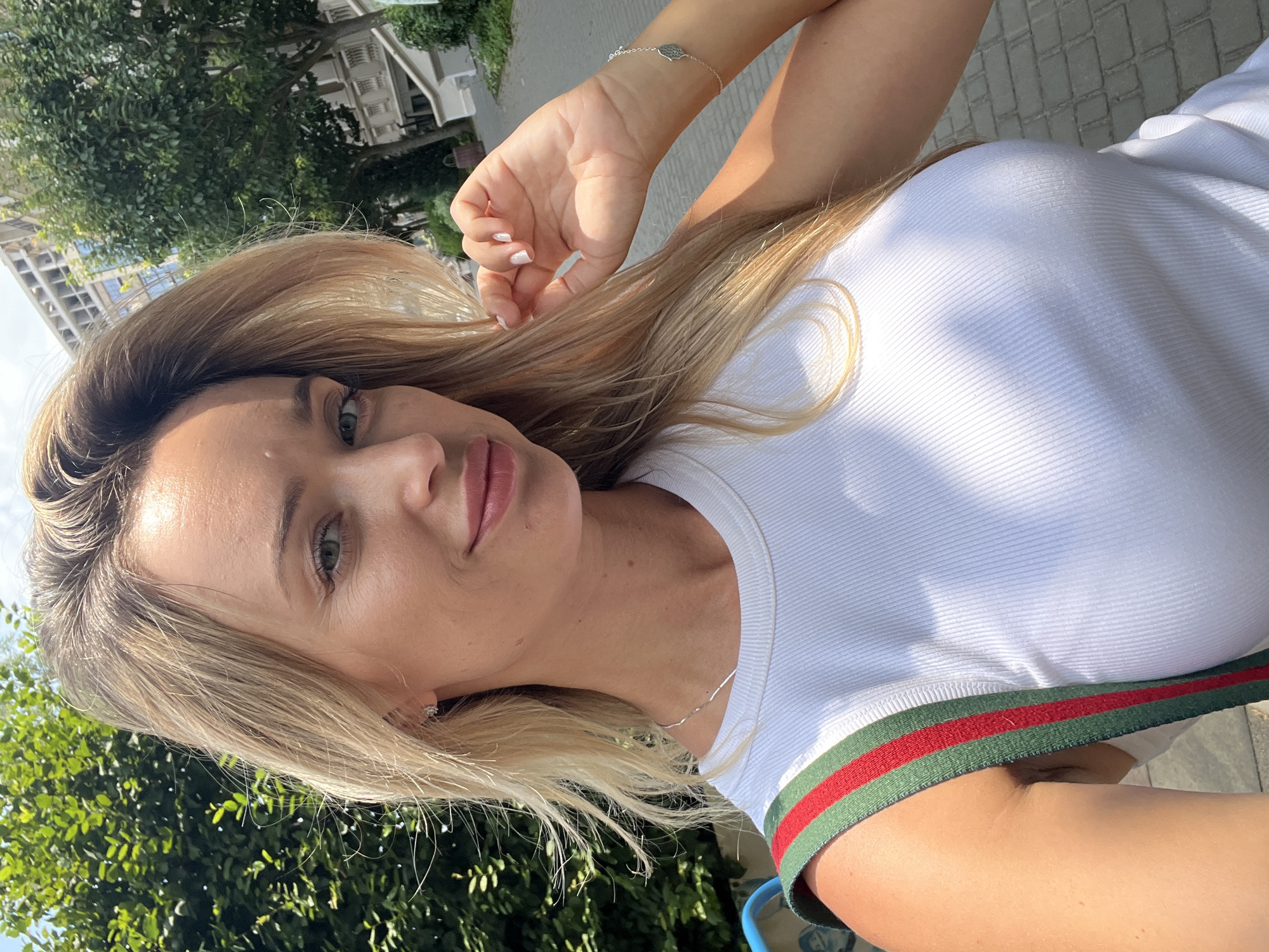 A young blonde woman is taking a selfie outdoors on a sunny day. She is wearing a white ribbed tank top with a green and red striped strap across her shoulder. She has her right hand raised, touching her hair near her face. She is wearing a delicate silver necklace and small stud earrings. A silver bracelet with a hamsa hand charm is visible on her right wrist. The background shows trees, buildings, and a paved walkway. The specific location is not discernible from the image.