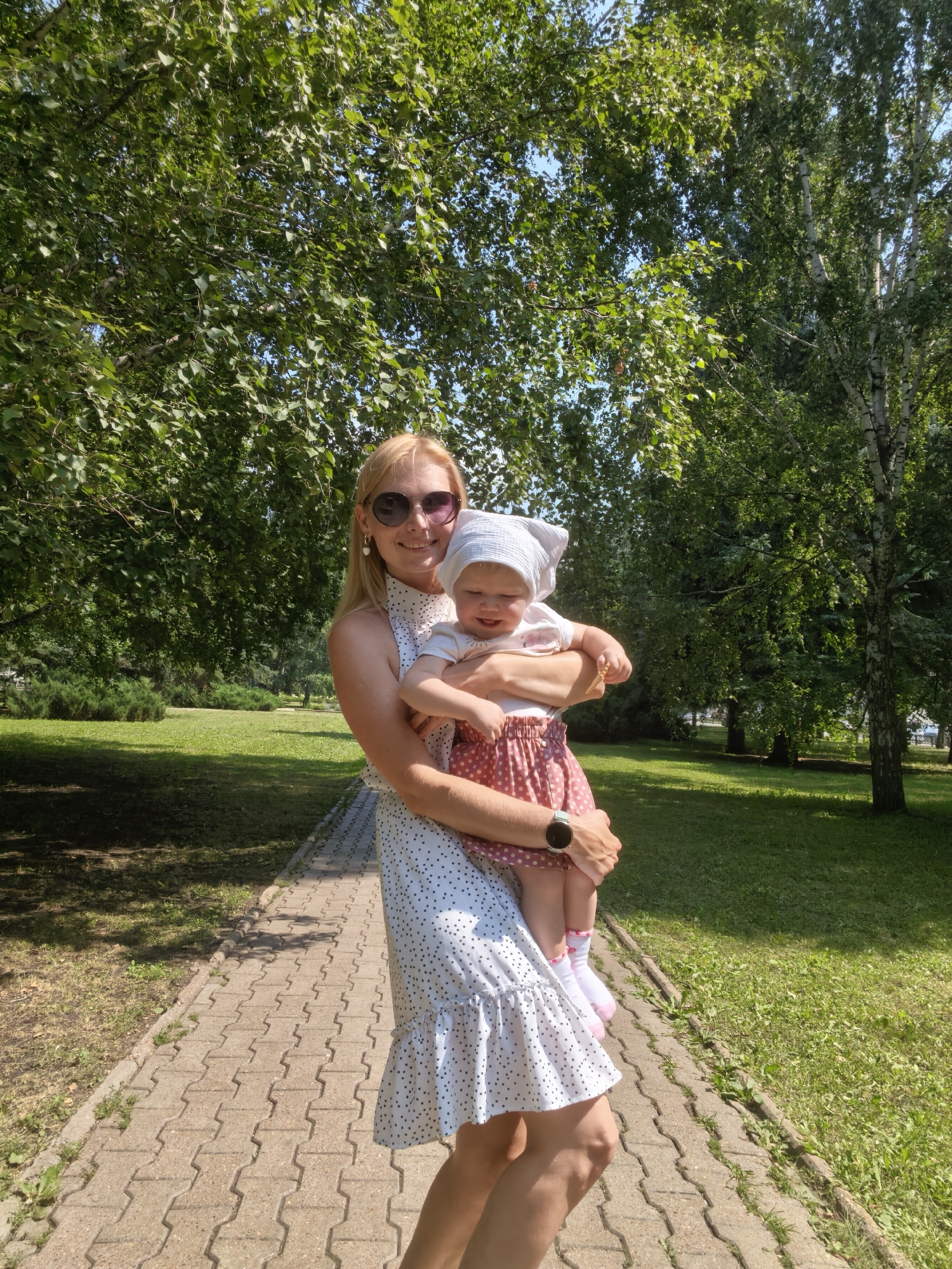 A blonde woman in sunglasses and a white polka dot dress holds a baby wearing a white head covering and a pink polka dot skirt. They are on a paved walkway in a park with lush green trees and grass. The woman is smiling. The baby is also smiling. The scene is brightly lit, suggesting it is daytime.