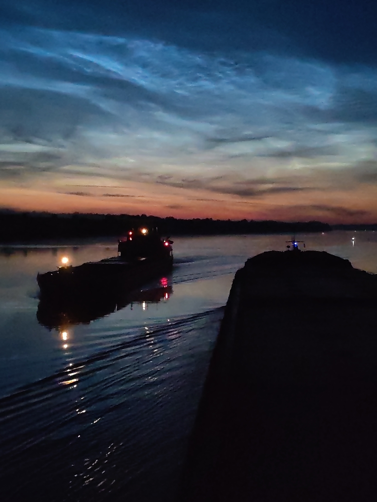 A barge with lights moves across calm water, reflecting the sunrise and the sky. The sky is filled with wispy clouds, tinged with orange and blue. The scene appears to be at dawn or dusk. The location is not identifiable as Ivanov Bor, Russia.
