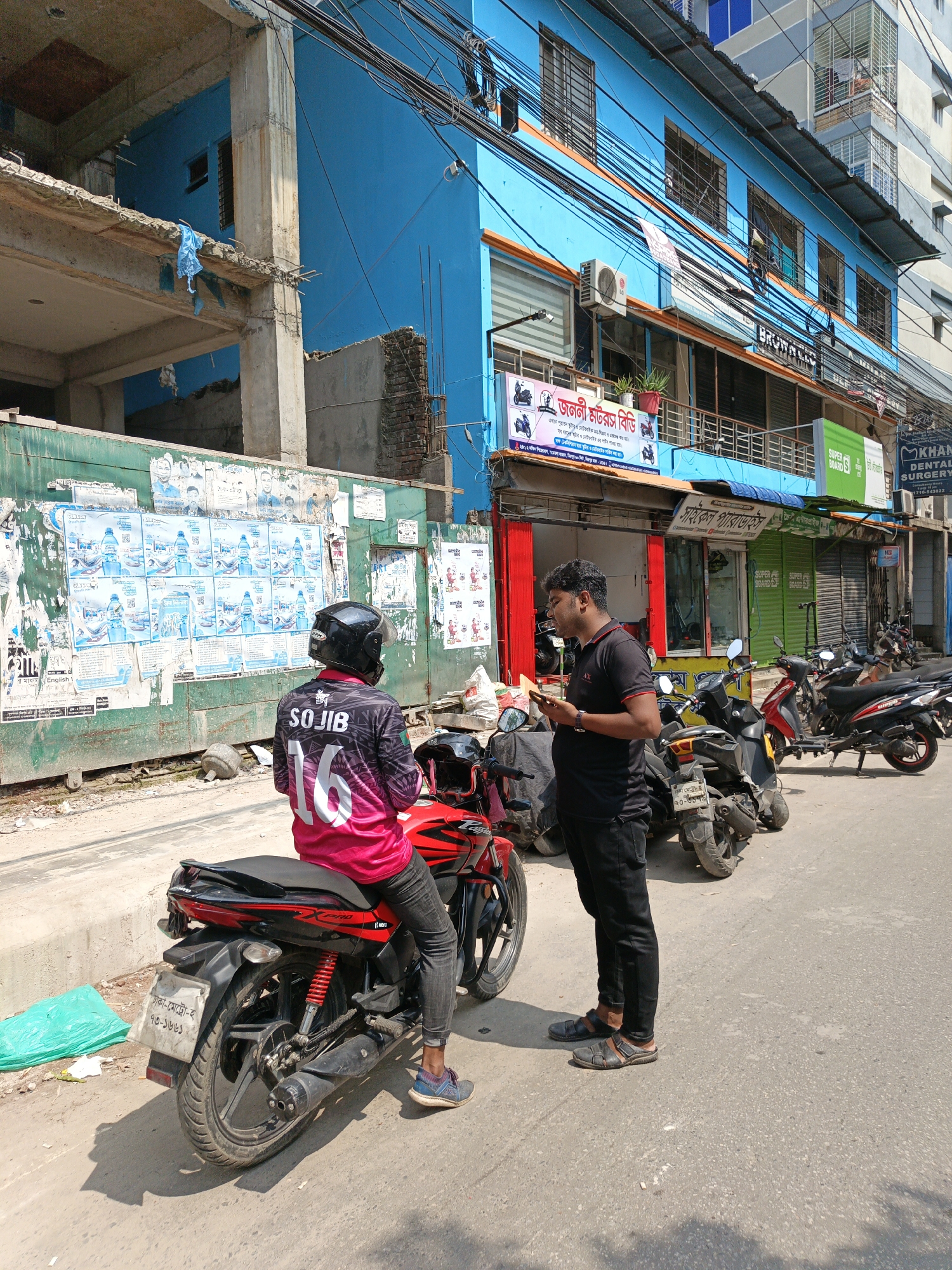 A street scene in Unknown, Unknown shows a man on a red and black motorcycle talking to another man. The motorcyclist is wearing a pink jacket with "SOJIB" and "16" printed on the back, jeans, and a helmet. The standing man is wearing a black shirt and pants and is looking at his phone. They are standing on a street lined with parked motorcycles and storefronts. The buildings are various colors, including blue, green, and red. A building under construction is also visible. Posters are attached to a green wall.