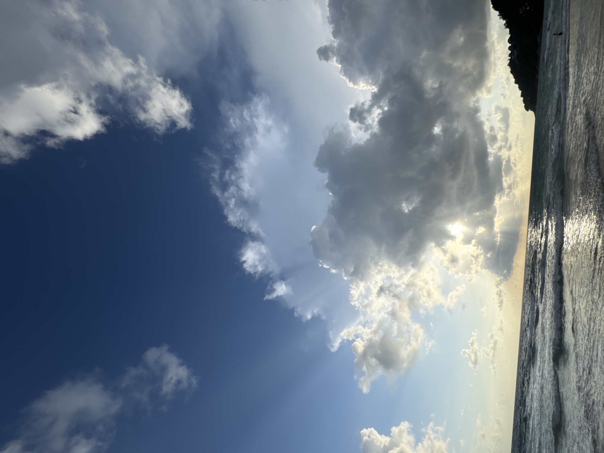 The image captures a dynamic sky over the ocean at Karon Beach, Thailand. The sky is a mix of deep blue and dramatic clouds. Sunlight pierces through a large, dark cloud formation on the right, creating visible rays that extend towards the left. Smaller, lighter clouds are scattered across the sky, with some appearing to be dissipating. The ocean is visible in the lower portion of the frame, with gentle waves breaking on the shore. The water reflects the sunlight, creating a shimmering effect. On the far right, a dark, tree-covered headland juts out into the sea. A solitary figure is visible standing in the shallow water near the shore, silhouetted against the bright light. The overall scene suggests a late afternoon or early evening, with the sun setting or about to set behind the clouds.