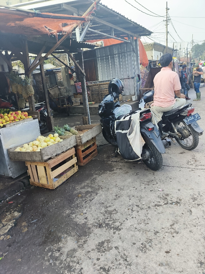 The image shows a street scene in Bandung, Indonesia, located at -6.906 latitude and 107.644 longitude. There are two motorcycles parked in front of a small, open-air market stall. One motorcycle is black, with a black helmet and bag, and the other is white, with a black seat and a black license plate that reads “D 3454 DC”. A man in a pink shirt sits on the white motorcycle, and another man in a blue shirt stands nearby. There are crates of fruit, including pineapples and yellow fruit, on the ground near the stall. The street is paved with gray concrete and there are some wires overhead. The scene appears to be in a busy commercial area.