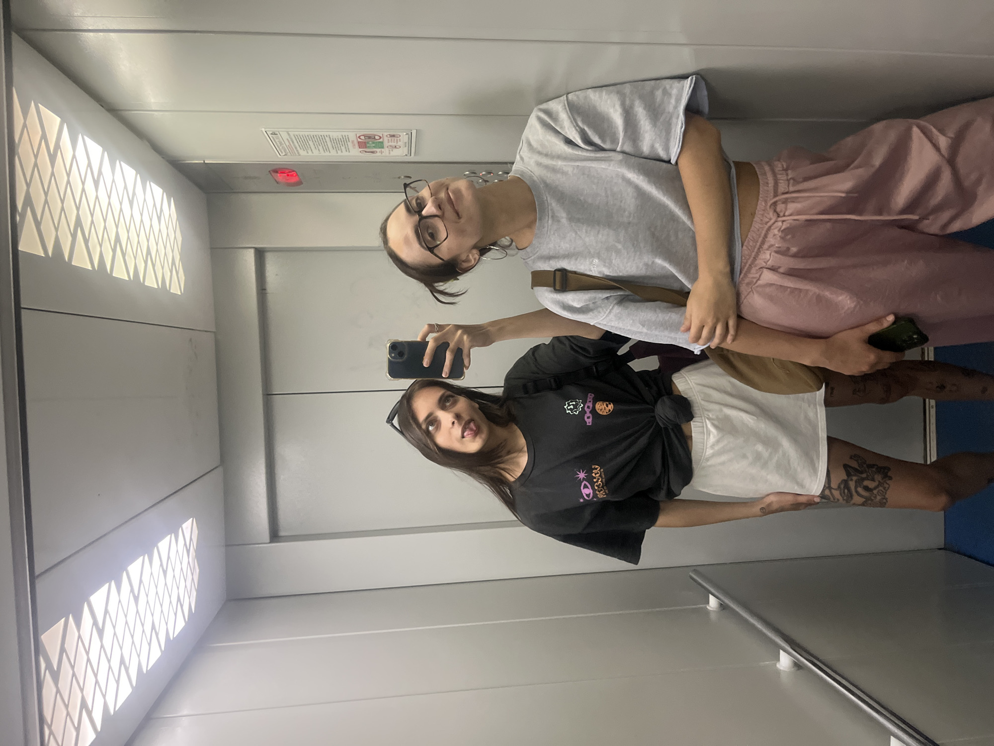Two young women are taking a selfie in an elevator. The woman on the left is wearing a black t-shirt tied at the waist, a white skirt, and sunglasses on her head. She is sticking her tongue out at the camera. The woman on the right is wearing a grey sweatshirt and pink pants, with glasses and large hoop earrings. Both women appear to be in their late teens or early twenties. The elevator has metal walls and a panel of square lights on the ceiling. There is a red illuminated button on the wall, and a sign with safety instructions. The floor of the elevator is blue. The image does not provide any information about the location of Krasnoyarsk, Russia.