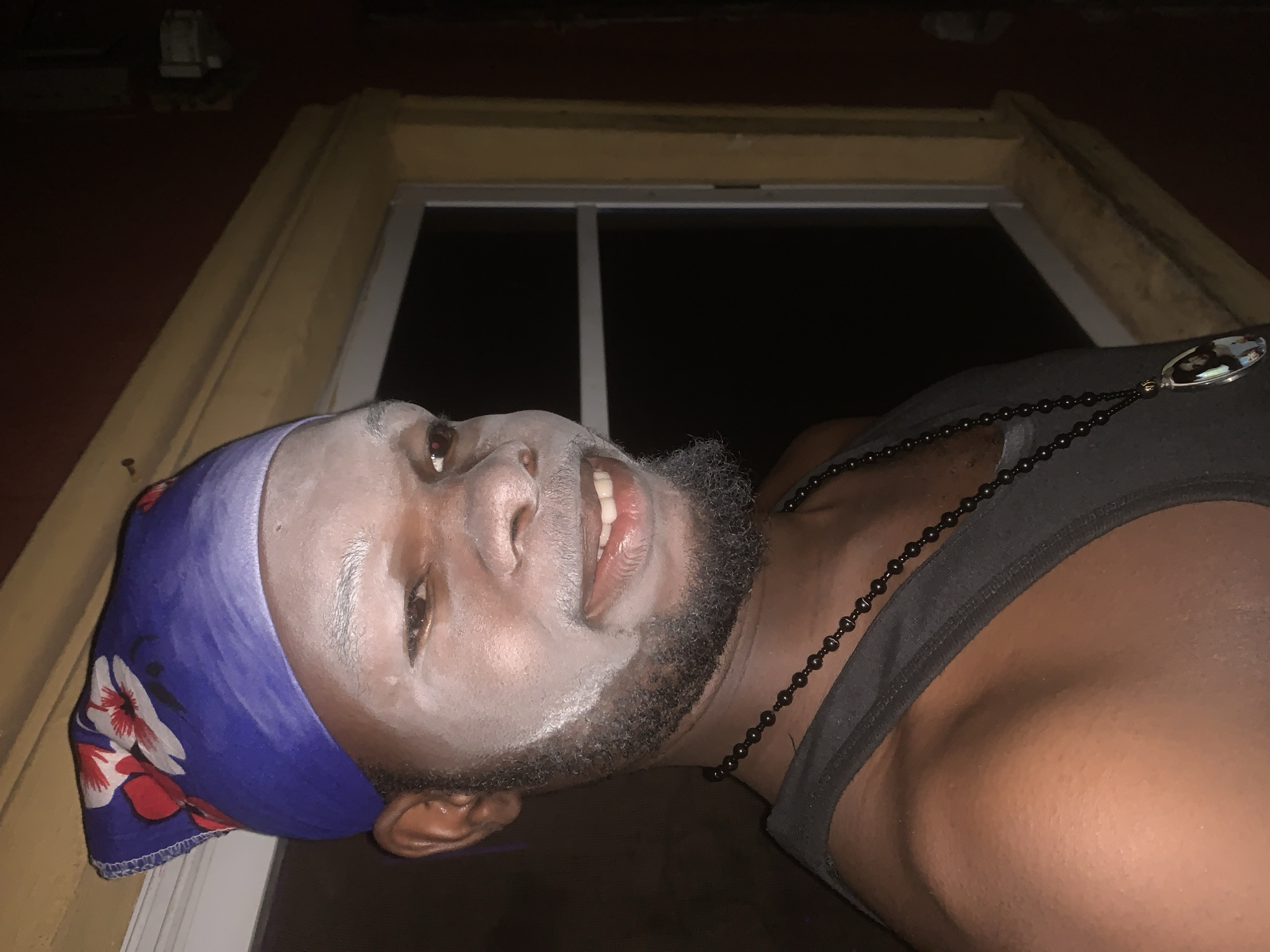 A low-angle, close-up shot captures a person of color, presumably male, indoors in Obosi, Nigeria, at night. The individual's face is largely covered in a light-colored, possibly white, powder or paint, leaving only the dark beard and portions of the skin visible around the eyes and mouth. He wears a blue headscarf adorned with white and red floral patterns. His expression is calm, with a slight smile, and he looks upwards and to the right.
He is wearing a dark, possibly black or grey, tank top and a black beaded necklace with a shiny, ornate pendant hanging on his chest. The background features a window with a white frame, beyond which is darkness, indicating the time of day is night. Around the window, there are wooden structures or frames, likely part of the building's interior, in yellowish and darker brown tones. The lighting in the image is dim, suggesting an indoor setting with limited illumination. There is no visible text in the image.