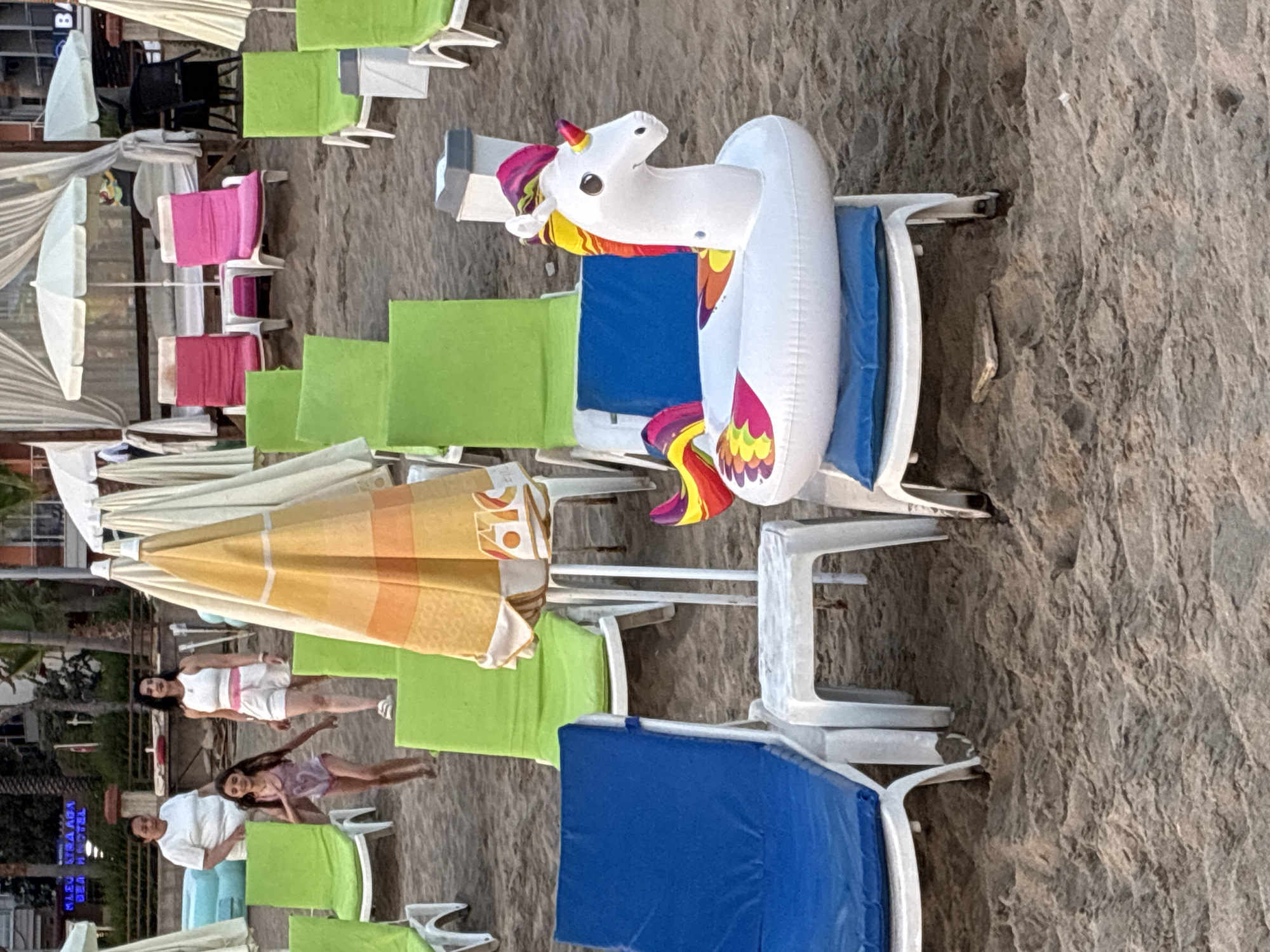 This is a photograph of a beach in Alanya, Turkey, with a focus on beach chairs and an inflatable unicorn. The scene appears to be set up for relaxation, with multiple rows of white beach chairs, some of which are equipped with bright green and blue cushions. Several beach umbrellas are also visible, with one prominently displaying yellow and orange striped fabric.
The most striking object is a large, white inflatable unicorn ring, positioned on one of the beach chairs. It features a colorful mane and tail with rainbow hues.
In the foreground, three people are visible. One person, wearing a white t-shirt and blue shorts, is standing. Another person, a woman in a white top and shorts with a pink waistband, is standing and looking towards the camera. A third person, a woman in a light purple patterned dress, is also standing. The presence of people suggests the beach is in use or being prepared for visitors.
The lighting and shadows suggest it might be late afternoon or early evening. The sand is visible throughout the image, indicating a sandy beach environment. The overall impression is a typical beach resort setting, likely in a popular tourist destination like Alanya, Turkey. No visible text is present in the image.