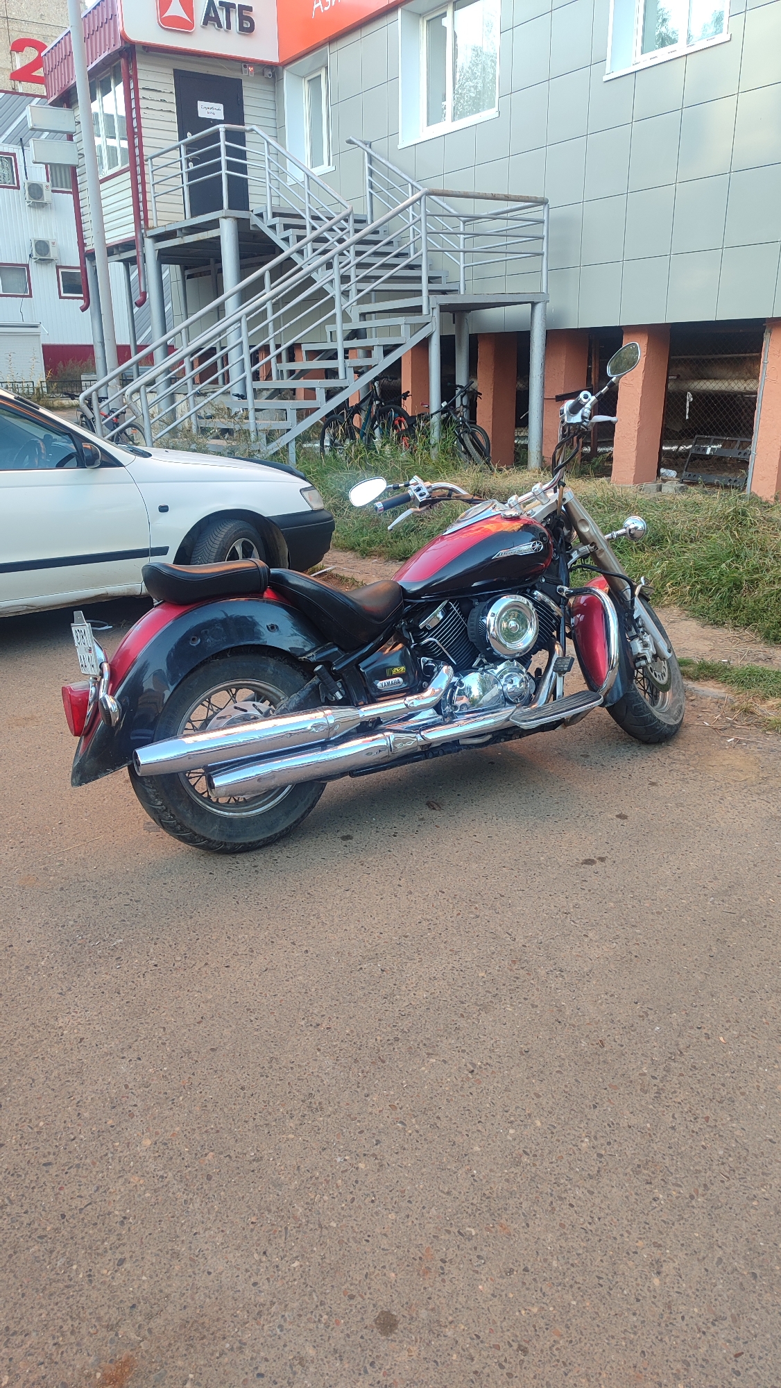 A black and red Yamaha motorcycle is parked on the asphalt in front of a building with a metal staircase. A white car is partially visible to the left of the motorcycle. The building has a sign with "АТБ" in red letters. In the background, several bicycles are leaning against the building. The scene appears to be in Mirny, Russia.