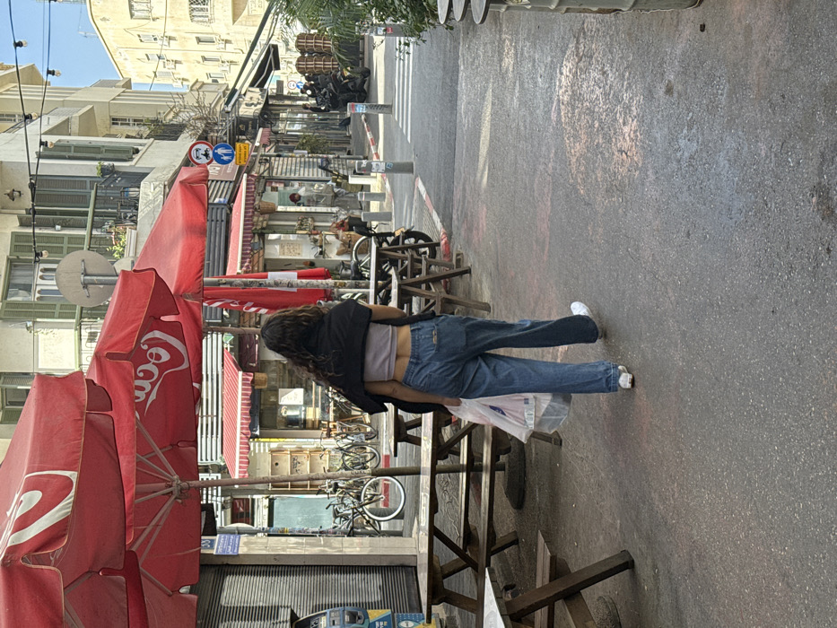 A daytime street-level image depicts an urban commercial area in Tel Aviv-Yafo, Israel. The central figure is a female, viewed from behind, with long, dark, wavy hair. She wears a light purple crop top, loose-fitting blue jeans, and white sneakers, with a dark jacket draped over her shoulders. She is walking away from the camera, carrying a clear plastic bag containing white and red items in her left hand.
The left side of the paved street features several wooden picnic tables with benches, positioned under three large red umbrellas, two of which display the white "Coca-Cola" logo. Behind these, multiple bicycles are parked against a building. A blue public utility box is visible on the far left. The adjacent building is multi-story, light-colored, and features balconies with closed green shutters and a satellite dish.
The street surface is dark gray asphalt with patches of reddish staining. Bollards delineate the path, leading towards a narrow passage where a white-striped crosswalk is visible. A street sign cluster includes a red 'no entry' symbol, a blue pedestrian crossing sign, and Hebrew text. Further into the background, parked motorcycles, indistinct figures, and additional buildings with various shopfronts and awnings are present, beneath overhead string lights.