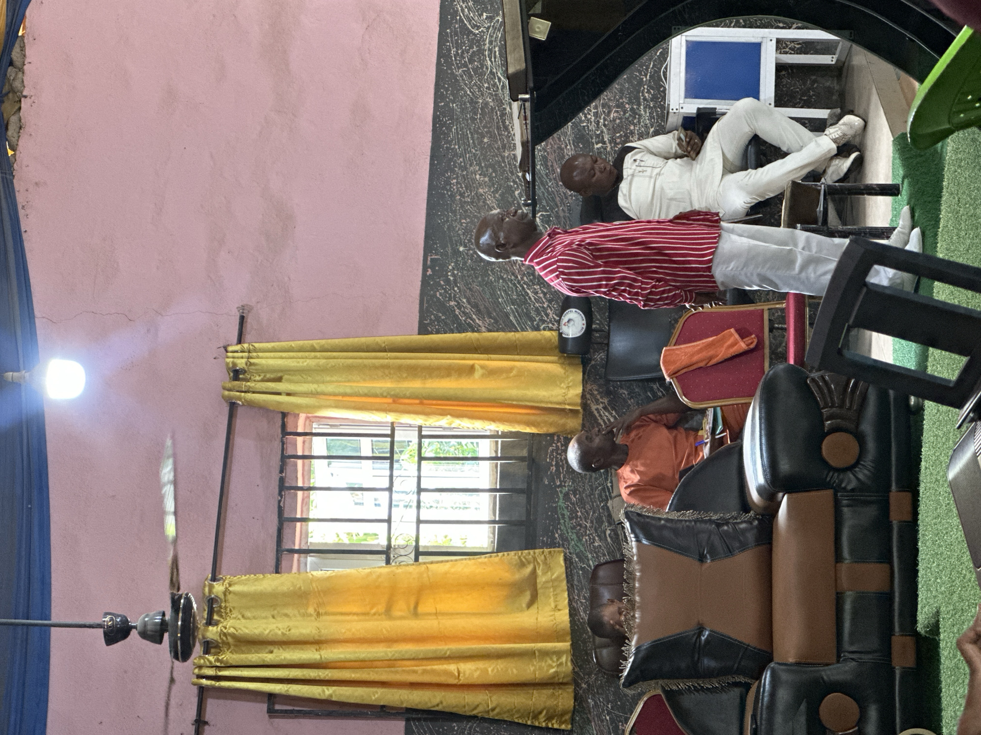The interior scene shows three men seated in what appears to be a room in Aba, Nigeria. A man in a red and white striped shirt and white trousers sits in the center of the frame, while another man in a white suit and shoes sits further back near what appears to be a staircase. A third man, wearing an orange shirt, is seated in the foreground.
A window with gold curtains is visible, allowing natural light into the room. A ceiling fan with rotating blades is positioned near the window. The room is decorated with patterned walls and includes various pieces of furniture, including chairs with leather cushions. There are objects, such as a bottle and a folded orange cloth, placed around the men. The floor is covered with a mix of materials, including a dark, swirling pattern and artificial green turf.