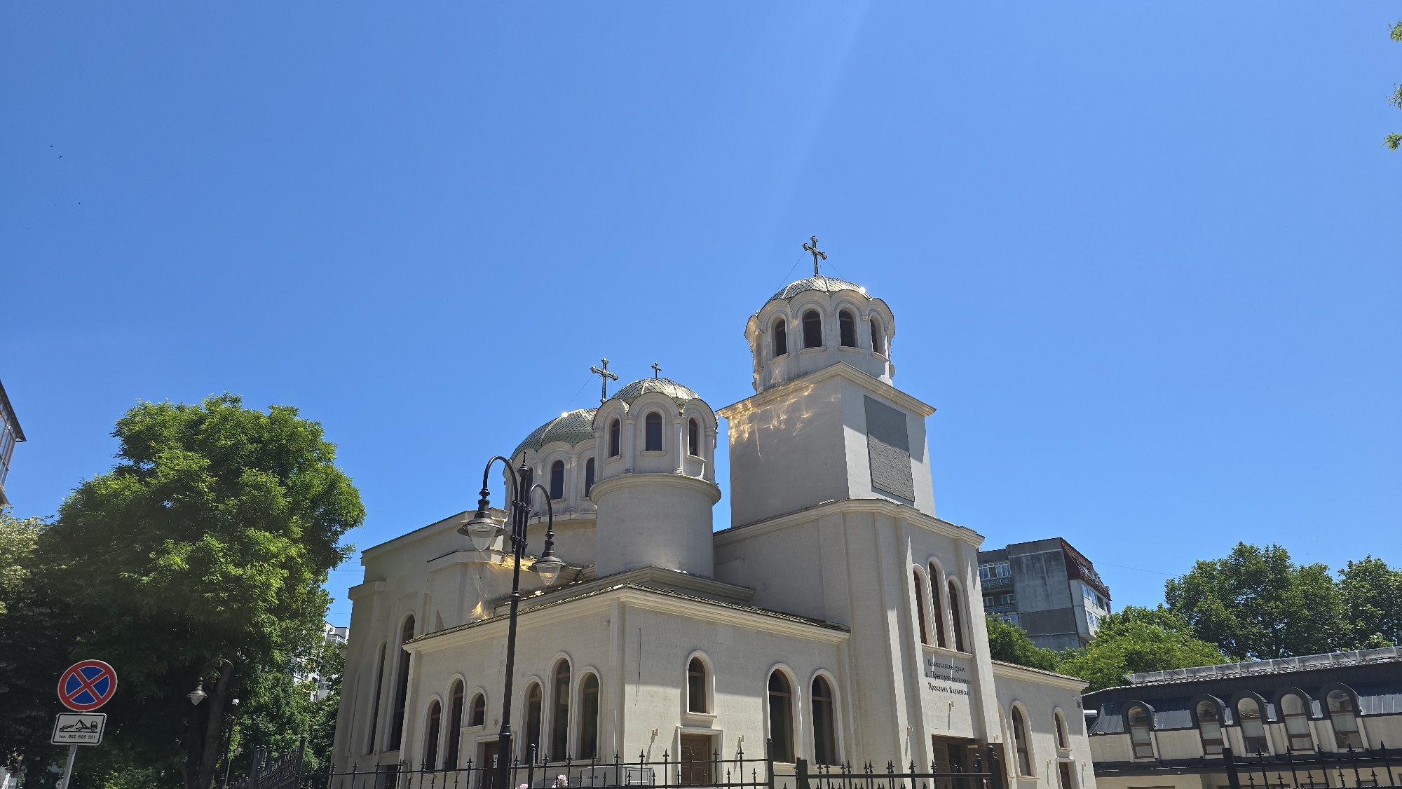 The image shows the Cathedral of the Assumption of the Virgin Mary in Varna, Bulgaria, on a clear, sunny day. The cathedral is a large, white Orthodox church with multiple domes, several of which are topped with golden crosses. The main dome is larger and more elaborate than the others, with an octagonal base and arched windows. A tall bell tower is attached to the right side of the church. The facade of the church features arched windows and decorative detailing. A street lamp with multiple lights stands in front of the church on the left side. In the background, there are tall green trees and several buildings, including a modern apartment building. A traffic sign is visible on the left, indicating parking restrictions. The sky is a vibrant, clear blue.
