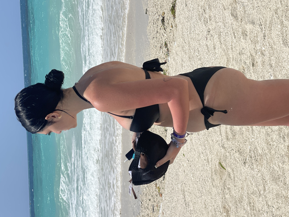 A young adult female is depicted standing in profile on a beach, facing left and looking towards the ocean, under a clear sky in Zanzibar, Tanzania. Her dark hair is styled in a bun. She is wearing a black two-piece bikini. In her left hand, held at waist level, she grips an inverted black mesh baseball cap containing a mobile phone and other small, indistinguishable items. Her left wrist displays multiple bracelets, including a dark blue band and several silver-toned ones. A small gold hoop earring is visible in her left ear, and a faint heart-shaped tattoo is present on her right upper arm. The visible skin on her arms, chest, and stomach exhibits a sun-kissed or slightly reddish tone. The foreground consists of light-colored sand and small, white shell fragments, with minimal green vegetation. The background features a vibrant turquoise ocean with moderate white waves breaking near the shore, transitioning to a deeper blue horizon under a bright, cloudless sky. No other individuals or man-made structures are discernible in the frame.