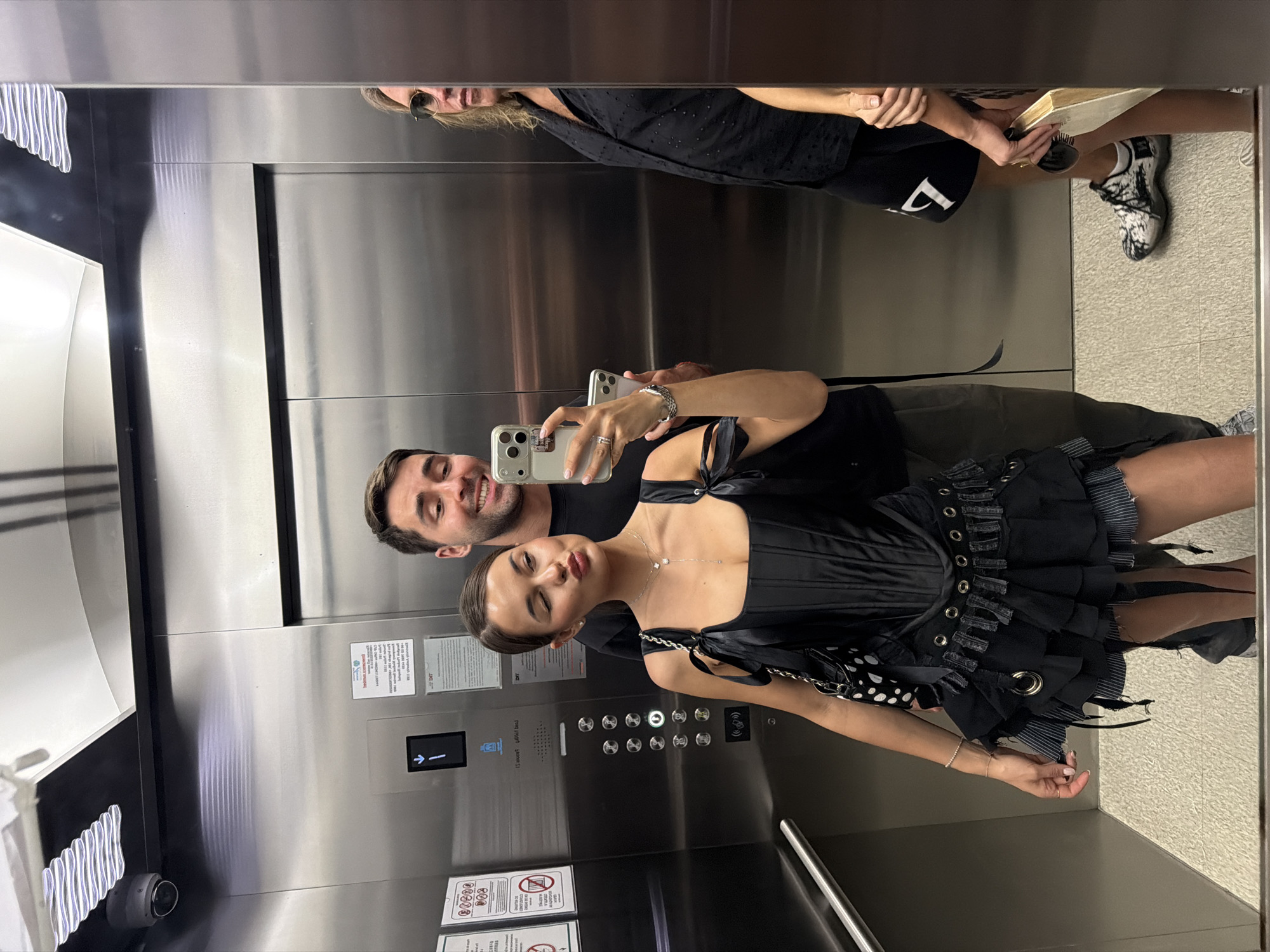 A man and a woman are taking a selfie in an elevator. The woman is in the foreground, wearing a black dress with a corset-like top and a ruffled skirt, and a pearl necklace. The man is behind her, holding the phone to take the photo. The elevator has stainless steel walls and an array of buttons. In the reflection at the top of the mirror, another woman is visible, wearing sunglasses and a black top, next to a pair of black and white patterned sneakers. There is a notice on the elevator wall. The location is Choeng Thale, Thailand.
