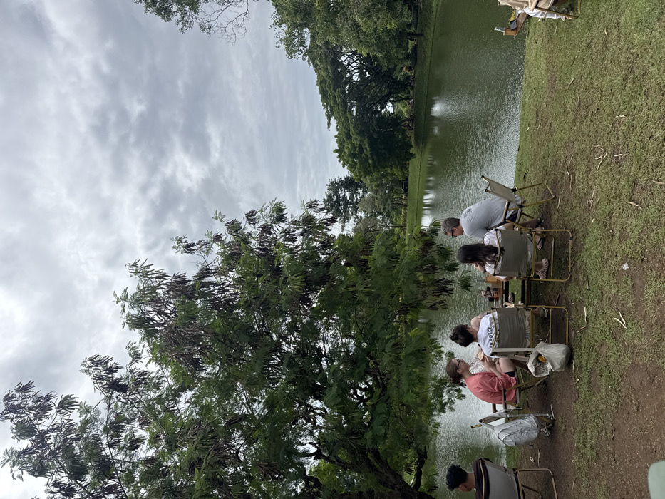 The image captures an outdoor gathering by a body of water on a cloudy day. The scene is oriented in landscape, depicting a grassy bank alongside a calm lake or large pond, with lush trees and an overcast sky.
At least five individuals are seated in portable folding chairs along the edge of the grassy bank, facing the water. From left to right in the main group of four:
A person wearing a light pink or coral-colored top is seated, facing slightly to their right. Next to them, a person in a white t-shirt with the word "LOVEST" clearly visible on the back is also seated, facing the water. A white plastic bag is placed on the ground near their chair. Further to the right, a person with dark hair and a light-colored top is seated. Adjacent to them, a man wearing a light gray t-shirt with sunglasses on his head is seated, looking towards the water or the people beside him. Small portable tables with drinks and other items are positioned in front of these groups. In the immediate foreground on the right, a fifth person with dark hair and a dark top is partially visible, also seated in a folding chair. In the far background, further down the bank, two additional figures are faintly seen seated.
A large, prominent tree with dense green foliage and numerous elongated, dark brown seed pods or dried leaves dominates the left side of the frame. The ground is covered in green grass, sloping gently towards the water. The body of water itself appears calm, reflecting the muted light of the sky and the surrounding trees, and is a dark green-brown color. The far bank of the water is lined with a dense growth of various green trees. The sky is entirely covered with light gray, diffused clouds, suggesting an overcast day with no direct sunlight. The activity appears to be a relaxed outdoor social gathering, possibly a picnic or a leisurely time spent by the water.