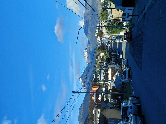 The image captures a daytime scene on a residential street in Ushuaia, Argentina, looking uphill towards a distant mountain range. The sky is largely clear blue with scattered white cumulus clouds. In the background, multiple mountains are visible, with several prominent peaks exhibiting snow cover.
The street itself is paved with asphalt, showing a darker, rectangular section in the mid-ground indicative of recent resurfacing or repair work. Vehicles are parked along both sides of the street. On the immediate left foreground, a light-colored minivan or car is partially visible, followed by a silver/light blue compact hatchback (consistent with a Citroën C3). Further up the street, a red SUV or crossover is parked on the right, near the resurfaced area, alongside other parked sedans and hatchbacks. No vehicles are in motion, and no occupants are visible.
Both sides of the street are lined with single- to two-story residential buildings of varying architectural styles and colors, including shades of yellow, beige, and brown. Overhead utility lines are prominent, strung between numerous wooden and metal utility poles, particularly on the left side of the frame. A streetlamp with a curved arm is positioned on the right side of the street in the mid-ground. Interspersed between the structures and along the roadside are various trees and shrubs. No individuals are depicted in the scene.