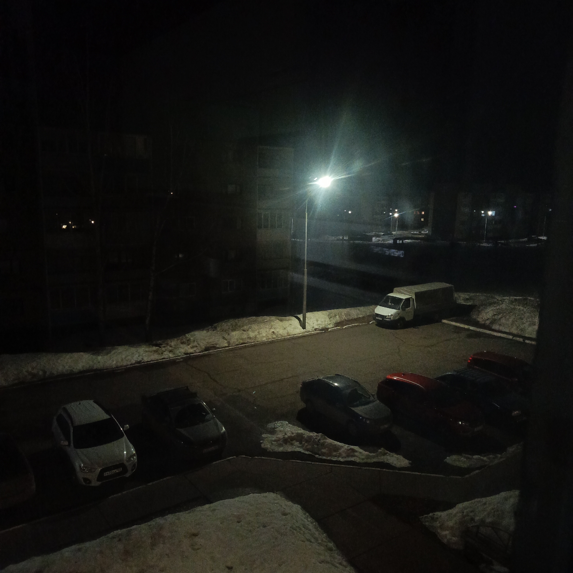 The image depicts a nighttime scene in what appears to be a residential area in Марковский, Russia. The view is from an elevated position, possibly from a building window. A parking lot illuminated by a streetlight is visible, containing several parked cars, including a white hatchback, a dark sedan, and a red vehicle. A white van is parked in the background near a snow-covered area. Buildings are visible in the background, shrouded in darkness, with few visible lights. Snow is present along the edges of the parking lot and on the ground in the surrounding areas. The overall lighting is dim except for the area directly illuminated by the streetlight. The presence of snow indicates a cold season. No people are visible in the scene.