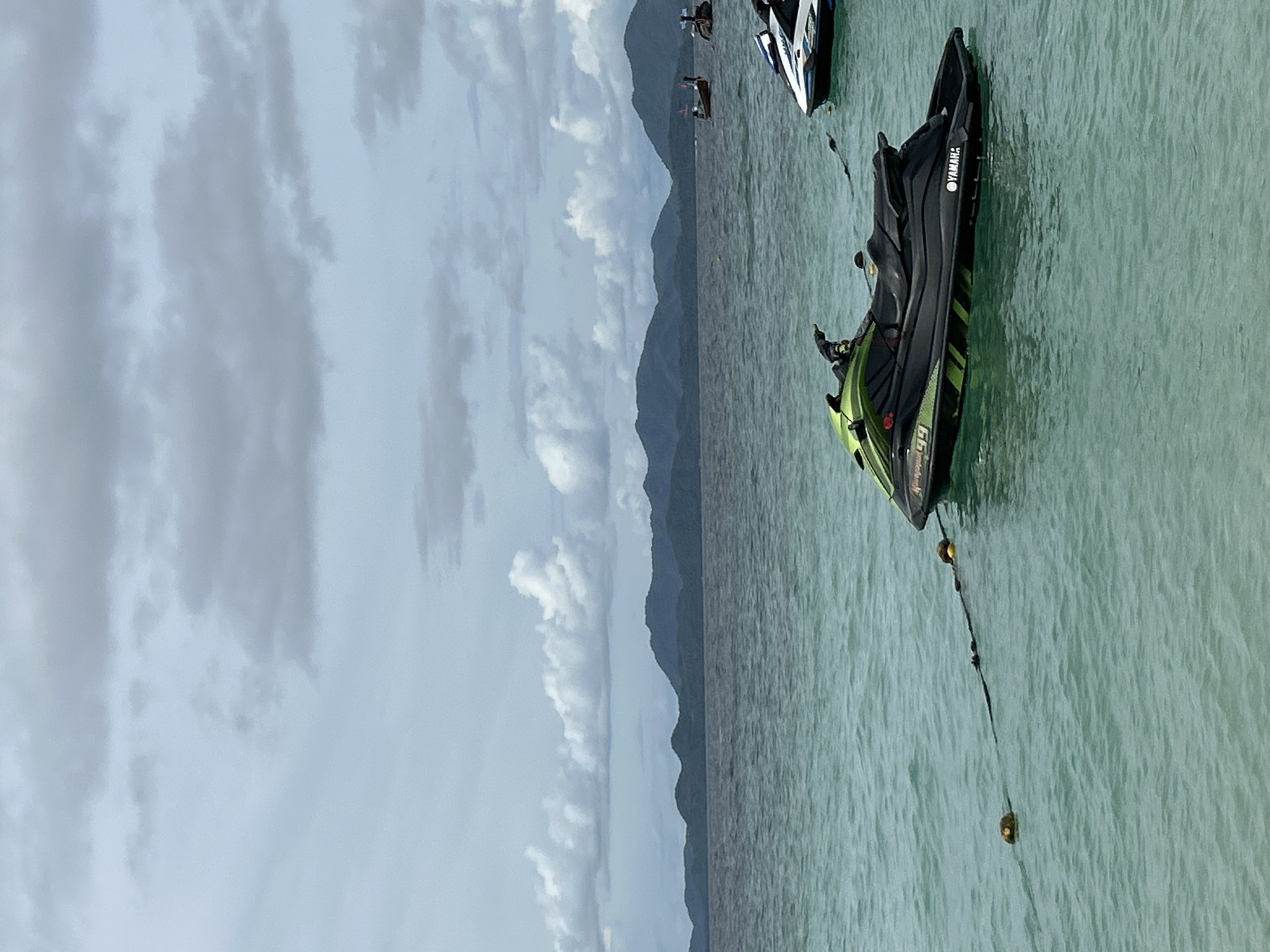 A tranquil scene unfolds on the ocean in Tambon Phru Nai, Thailand, with a cloudy sky overhead. In the foreground, a black and green Yamaha jet ski is moored to a buoy line, gently bobbing on the turquoise water. Further out, another jet ski, white with blue accents, is partially visible. In the middle ground, several traditional longtail boats are anchored, their slender forms silhouetted against the hazy backdrop of distant, verdant mountains. The calm sea reflects the overcast sky, creating a serene and slightly subdued atmosphere.