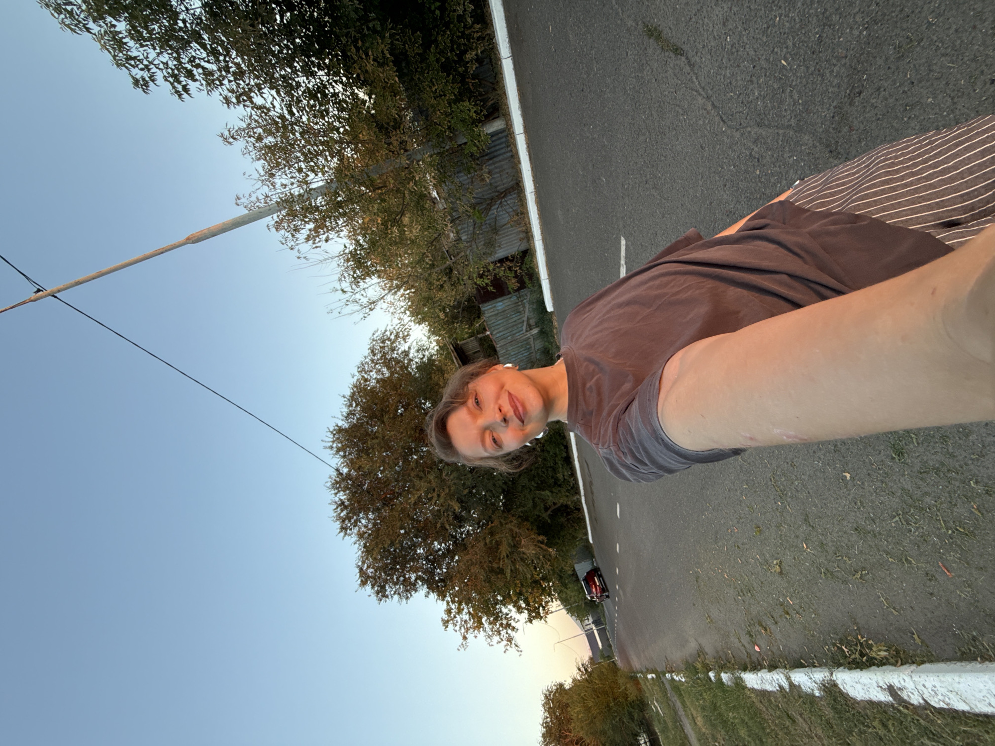 A woman is captured in a selfie on a paved road, likely in Aksay, Kazakhstan. The low angle of the shot emphasizes the sky, which transitions from a clear blue at the top to a warmer hue near the horizon, suggesting it's late afternoon or early evening. A tall utility pole with a wire stretching across it stands to the right of the frame.
The woman, with short, light brown hair and subtle makeup, is smiling at the camera. She wears a loose-fitting, dark grey t-shirt and striped pants. Her left arm is extended towards the camera, showing a patch of what appears to be dry, scaly skin on her forearm.
The road is asphalt with white dashed lines marking lanes. A white curb runs along the left side of the road, bordered by dry grass and some scattered debris. On the right side of the road, a weathered fence made of corrugated metal is visible behind a line of trees with autumn foliage. Further down the road, a red car is parked. The overall scene depicts a somewhat rural or suburban setting.