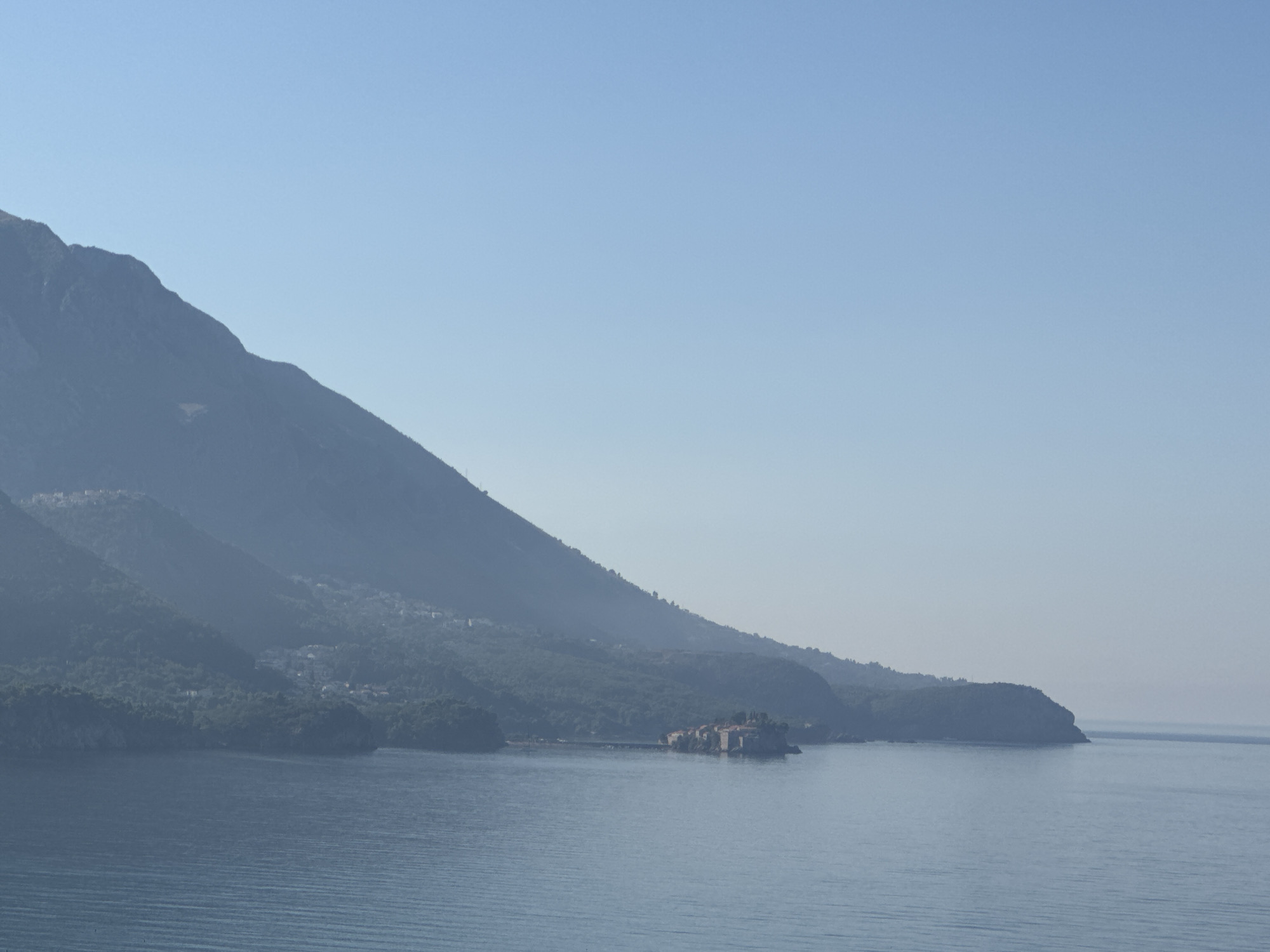 The image shows a view of the Adriatic Sea and the coastline of Boreti, Montenegro. A large, green, mountainous landscape dominates the left side of the frame, sloping down towards the sea. The lower slopes are covered in dense vegetation and dotted with what appear to be small buildings, suggesting a settlement or village. In the middle distance, a small island, known as Sveti Stefan, is visible jutting out into the sea. This island is connected to the mainland by a narrow causeway and is characterized by a cluster of old stone buildings with terracotta roofs. The sea itself is a calm, deep blue, with gentle ripples on its surface. The sky is a clear, pale blue, with no clouds visible. The overall atmosphere is serene and picturesque.
