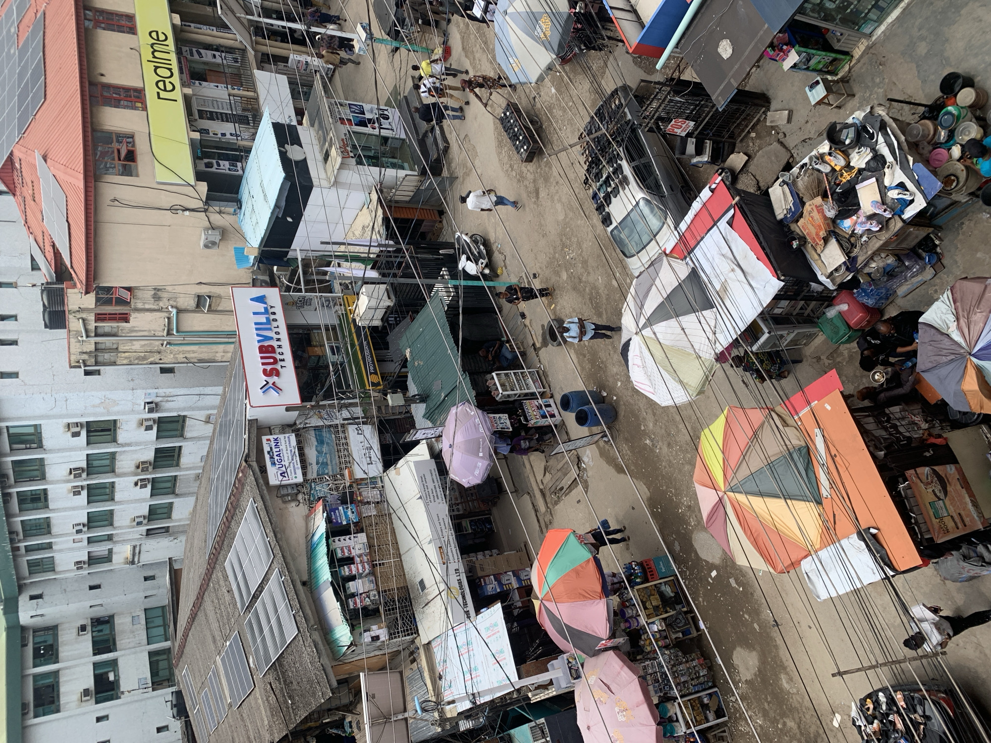 This is an aerial view of a street in Ikeja, Nigeria. The street is bustling with activity. People are walking and interacting with vendors under colorful umbrellas. The architecture consists of multi-story buildings with commercial signage, including brands like "realme" and "SUBVILLA TECHNOLOGY". Some buildings have solar panels installed on their roofs. Electrical wires crisscross above the street. Cars are parked along the street. One area includes what appears to be piles of used household items.