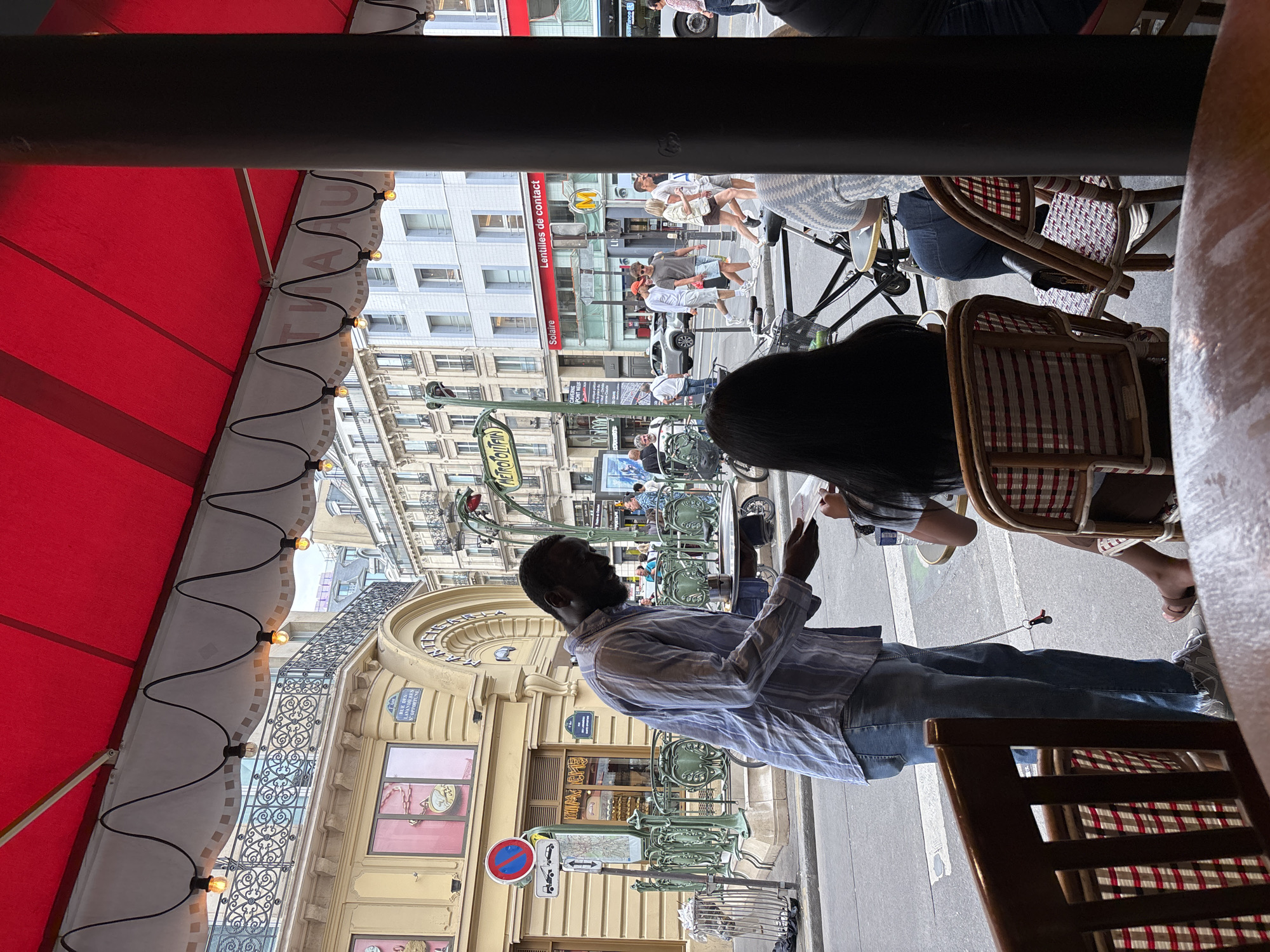 The image depicts a street scene in Paris, France, viewed from a cafe with a red awning. The perspective is rotated, making the scene appear sideways. In the foreground, a Black man wearing a blue and white striped shirt and jeans is seated at a table. He is holding a phone or tablet in his hands and looking upwards. To his right, a woman with long dark hair is seated, and beyond her, two chairs with red and white checkered cushions are visible.
The background shows a bustling street with people walking, cars, and iconic Parisian architecture. A green Metro entrance with the distinctive "Metropolitan" sign is prominent. Buildings with ornate balconies and shopfronts line the street. The lighting suggests it is daytime, possibly late morning or afternoon, with bright sunlight casting shadows. The overall atmosphere appears to be lively and typical of a Parisian outdoor cafe.
Visible text includes "Metropolitan" on the Metro sign, and "Solaire" and "Lentilles de contact" on a sign on a building. Another sign below the Metro entrance appears to be a street sign, but the text is not fully legible.