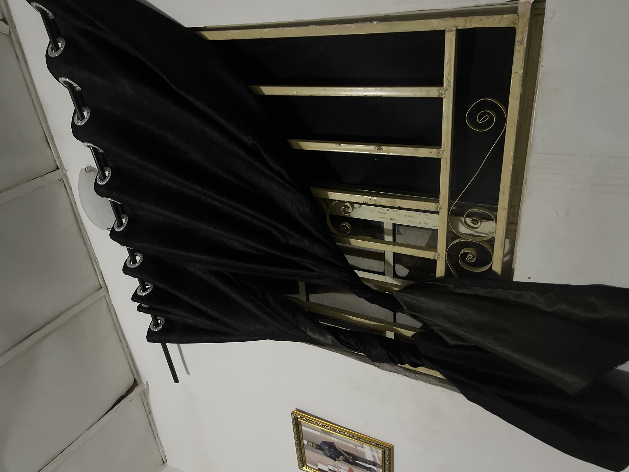 In a room in Howayi, Nigeria, black curtains with grommets are hung on a rod above a window. The window has a decorative gold metal grate with scrollwork. To the left of the window, a framed portrait of a man in a black suit is hanging on the wall. The ceiling appears to be made of white panels. The room is dimly lit, casting shadows on the walls and curtains.