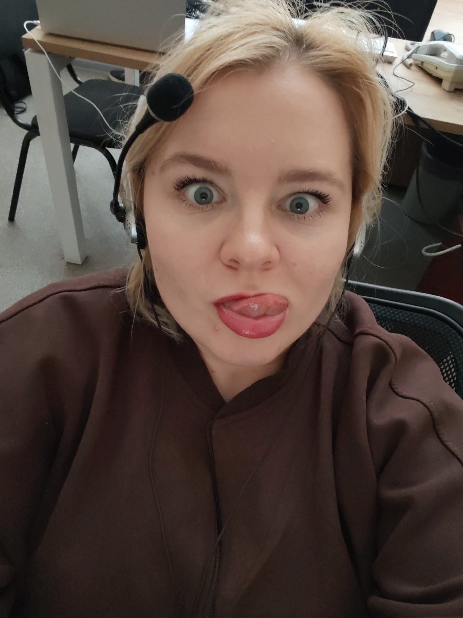 A blonde woman with blue eyes and wearing a headset is sticking her tongue out at the camera. She is wearing a brown top. The background includes office furniture such as desks, chairs, and a telephone. The location is likely an office setting, but there is no information about Izhevsk, Russia, visible in the image.