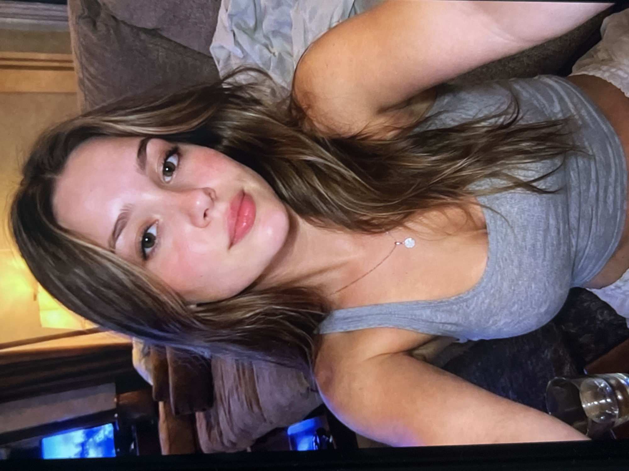 A young woman with long, light brown hair and brown eyes is captured in a close-up, head-and-shoulders shot. She is wearing a grey tank top and a delicate silver necklace with a small, round pendant. Her skin has a warm, tanned tone, and her lips are adorned with a glossy, pink lipstick. The background reveals a dimly lit room with a couch covered in a light-colored throw and a television screen displaying a bright, blue image. The overall ambiance suggests a casual, indoor setting.