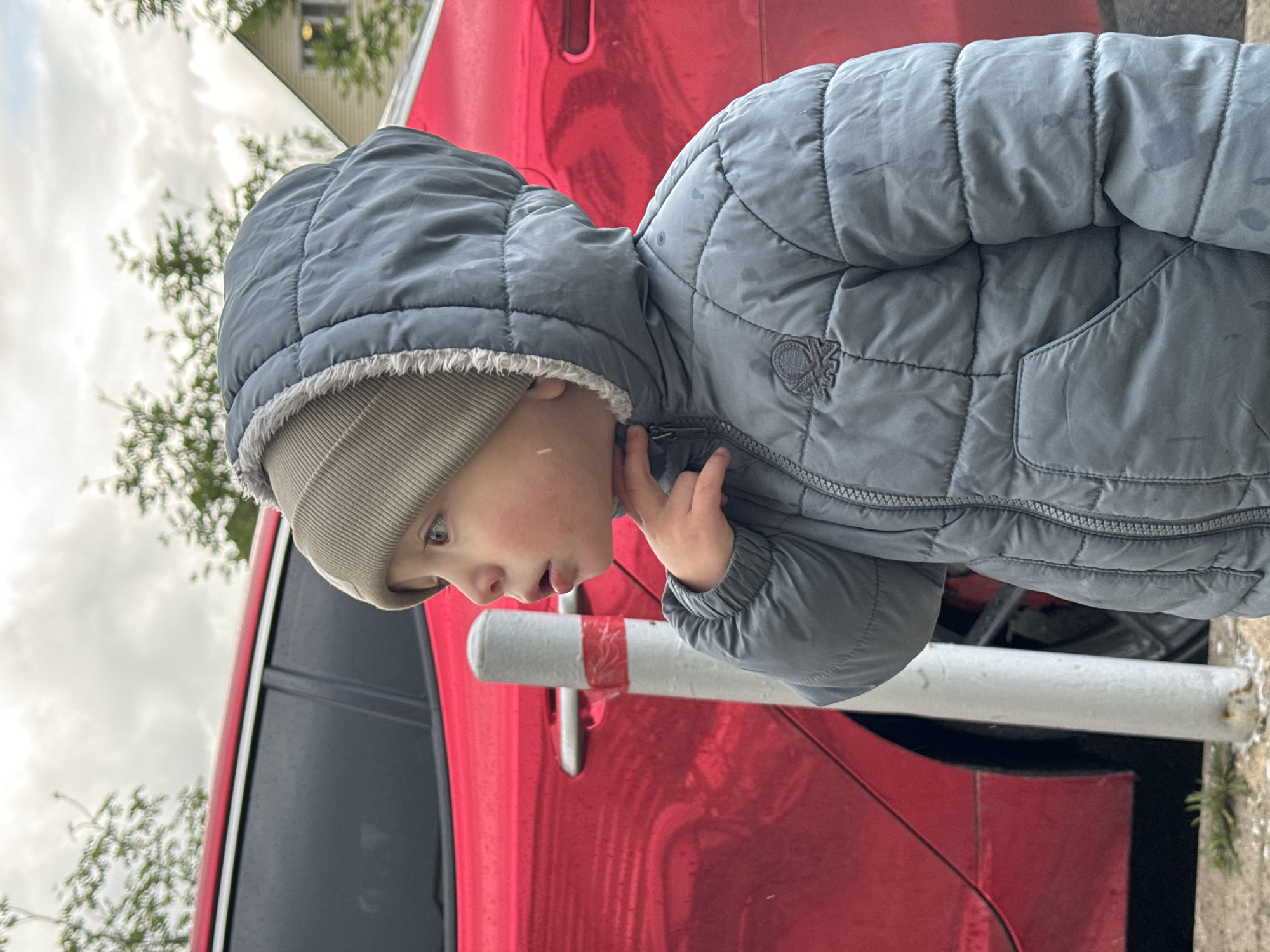 A young child, wearing a gray quilted jacket with the hood up and a gray beanie, stands near a red car. The child's right hand is near their face. A white and red striped pole is also visible. The scene appears to be outdoors, with trees and a building in the background. The location is identified as Akulovo (Gp Kubinka), Russia.