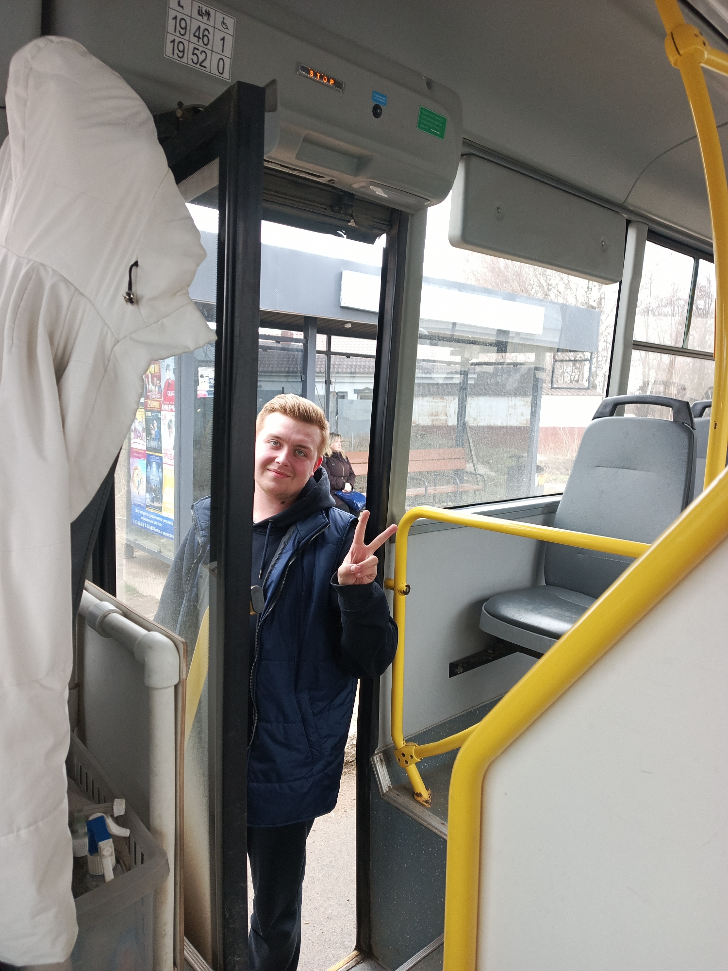 The interior of a bus in the city of Chaikovsky, Russia, is shown in a medium shot. A young man with fair skin and reddish-blonde hair is standing in the doorway, partially obscured by the door frame. He is wearing a dark blue vest over a black hooded sweatshirt and dark pants, and he is making a peace sign with his right hand. Outside the bus, visible through the glass doors, is a bus shelter with posters. A woman is sitting on a bench in the shelter. Inside the bus, a white hooded coat hangs to the left of the doorway. A yellow safety bar is on the right side of the doorway. Gray bus seats are visible. A small container with cleaning spray bottles is visible near the coat. A sign above the door reads "19 46 1" and "19 52 0." A "STOP" sign is illuminated above the door. The bus appears to be at a stop.