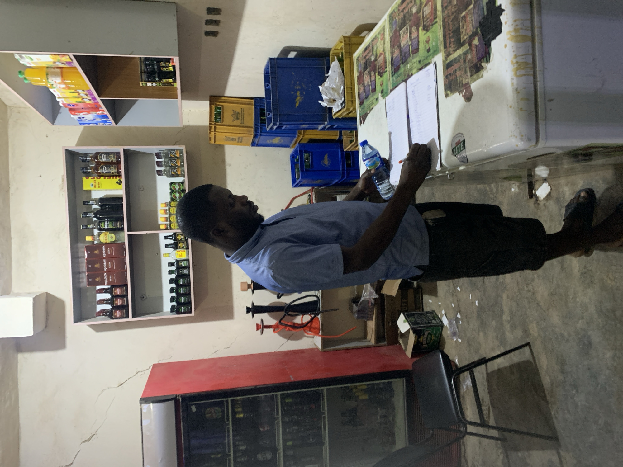 A man stands in a room that appears to be a small business or convenience store in New Stadium, Nigeria. He is writing on papers affixed to a refrigerator. He is holding a water bottle in one hand and a pen in the other. The room contains shelves stocked with beverages, including bottled juices and liquor. Blue and yellow crates are stacked nearby. A red cooler is also visible. The floor is concrete.