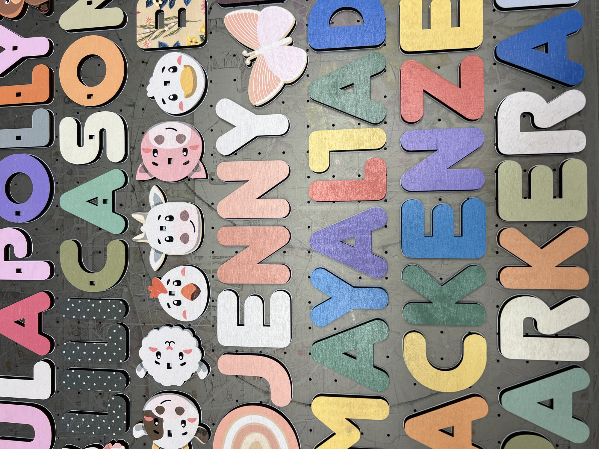 This is a close-up, overhead shot of a collection of colorful wooden letter and animal cutouts arranged on a dark gray, textured surface. The letters are of various colors, including orange, purple, green, blue, yellow, pink, and white, and appear to be in a grid-like pattern. Some letters have a wood grain texture with a floral pattern, and others have a subtle metallic sheen. There are also several animal cutouts: a butterfly with pink and white wings, a white sheep with a black face, a white cow with brown spots and a pink nose, and a pink pig-like creature. The letters and animal cutouts are all three-dimensional with black edges. The surface they are on has a speckled texture and appears to be a bulletin board or similar material with small pinholes. The lighting is bright and even, casting subtle shadows that highlight the depth of the cutouts. No specific time of day or weather is indicated. The overall impression is a colorful and playful arrangement, possibly for a craft project, educational display, or decoration.