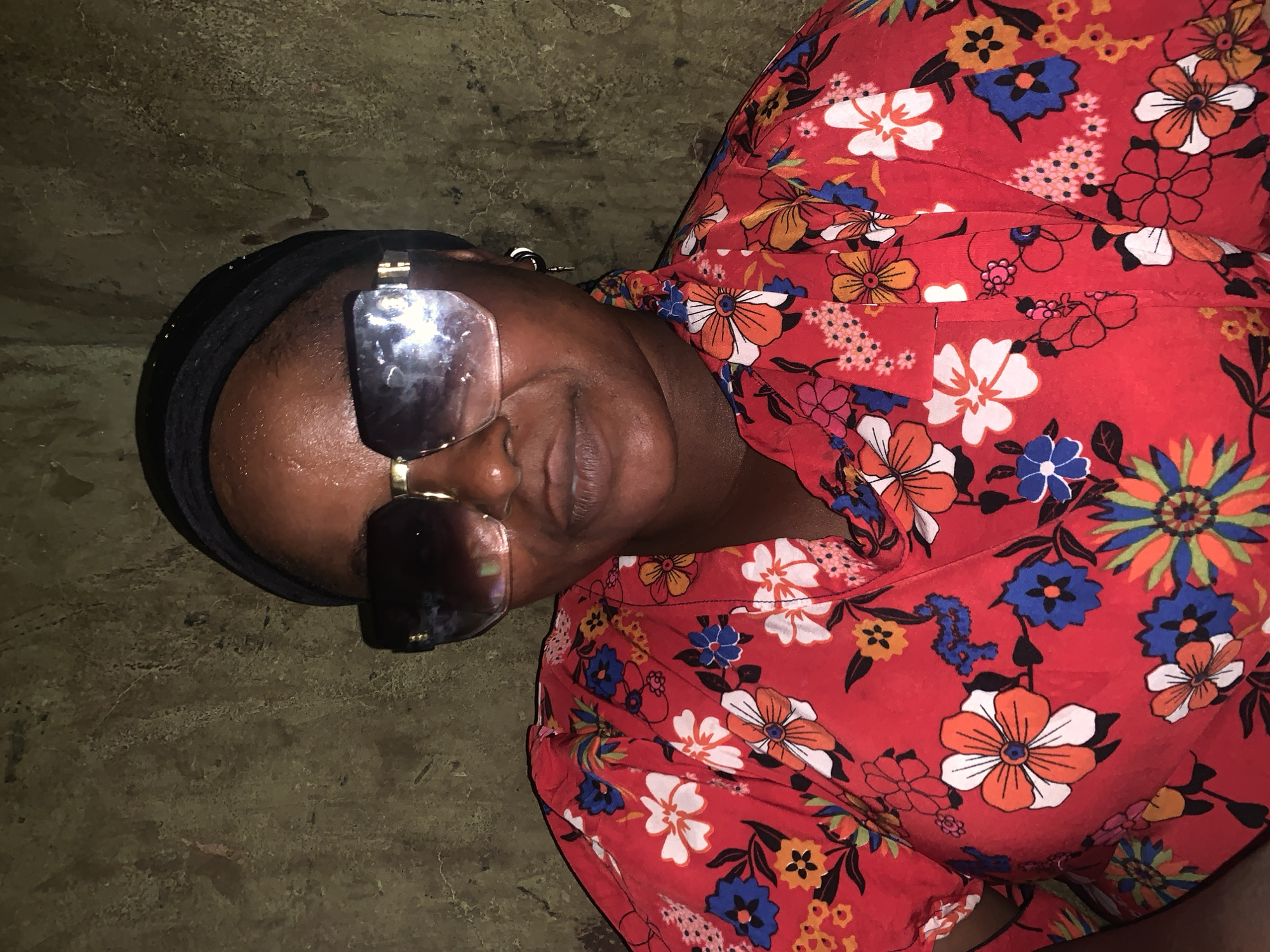 The image shows a person lying down, wearing a red dress with a floral pattern. The person is also wearing sunglasses and a black head covering. The background appears to be a textured, earthy surface, possibly a wall or floor. The lighting is dim, suggesting an indoor setting or a scene taken at night. The person's face is visible, but the sunglasses obscure their eyes. Their expression is neutral. The overall scene is still and lacks any overt action. There is no visible text in the image. The context provided indicates the location is Boi A, Nigeria.