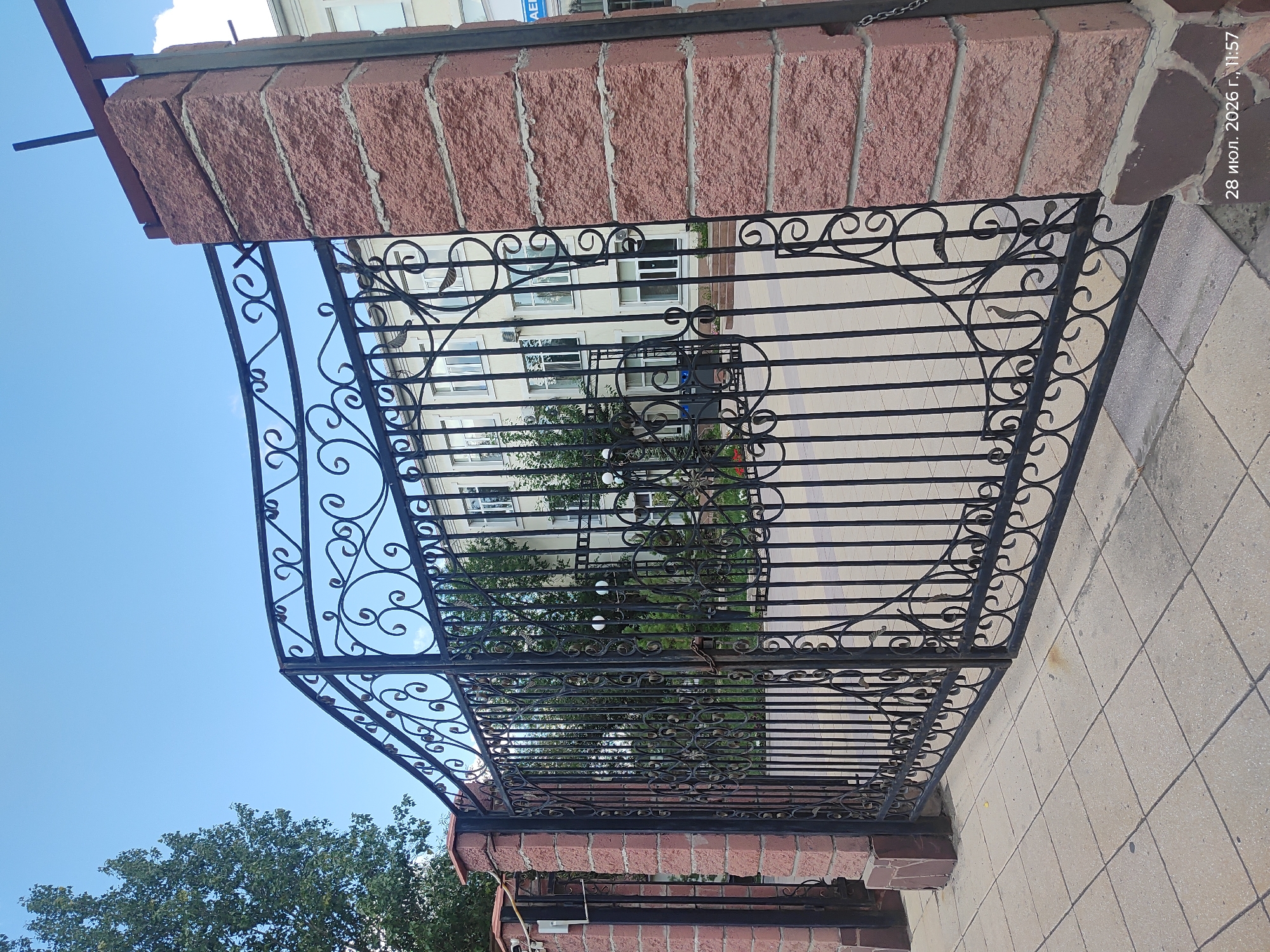 The image shows a large, ornate black wrought iron gate in front of a light-colored building with several windows. The gate is supported by two thick pillars constructed from rough-cut reddish-brown stones. The sky above is clear and blue with a few wispy white clouds. To the left of the gate, a leafy green tree is visible. The ground in front of the gate is paved with square light-colored tiles. The building in the background appears to be an educational institution or administrative building, given its multi-story structure and uniform windows. The timestamp in the lower right corner of the image indicates the photograph was taken on July 28, 2026, at 11:57 AM. While the image quality is good, no people are clearly visible within the frame. The overall impression is one of a well-maintained entrance to a public or institutional space.