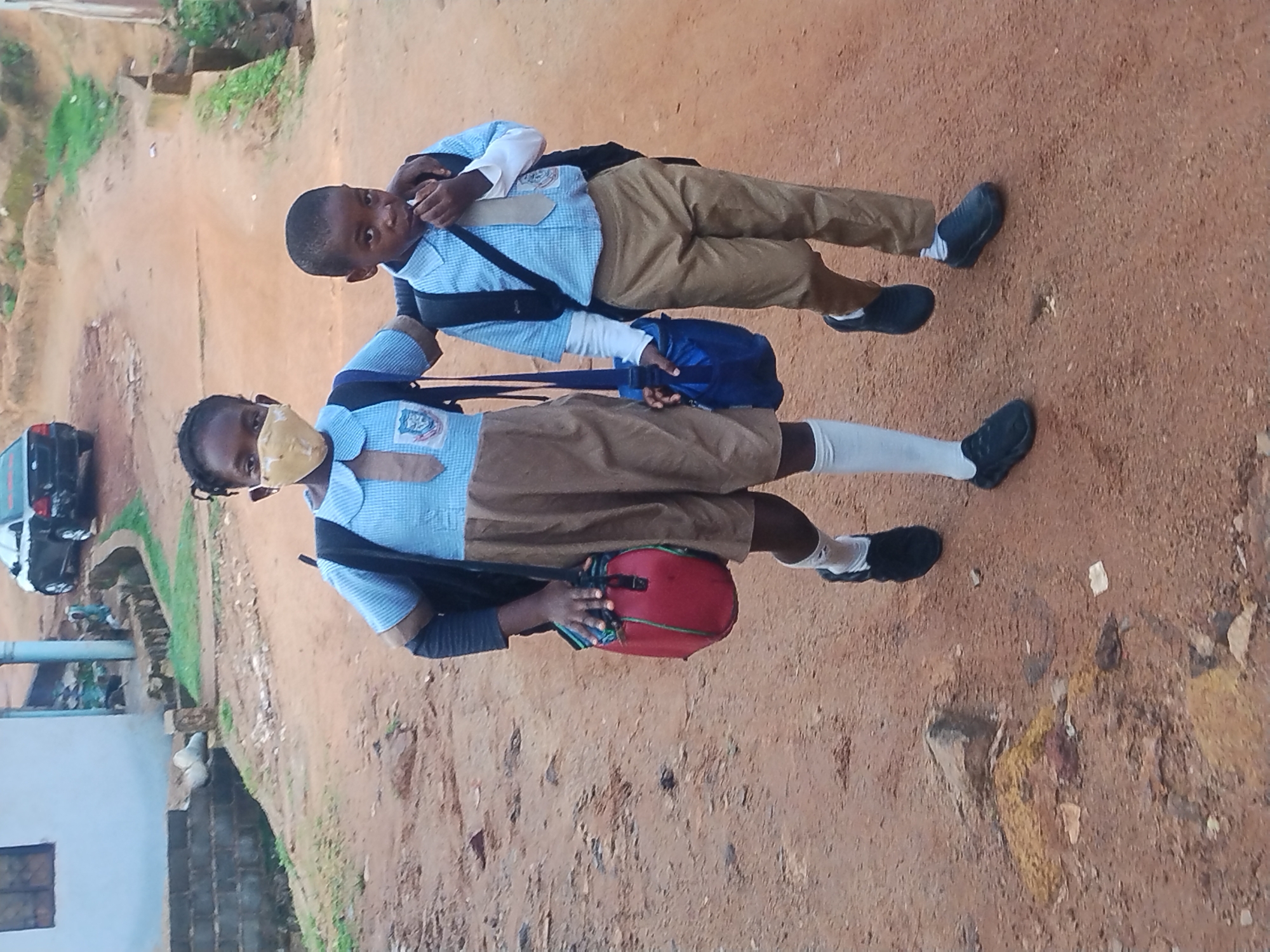 Two young children, a girl and a boy, walk along a dirt path in an urban environment, possibly on their way to or from school. The girl, on the left, wears a blue and tan school uniform, a light yellow face mask, and carries a red cylindrical bag and a black backpack. The boy, on the right, also in a blue and tan school uniform, has his hand near his mouth and carries a blue bag and a black backpack. Both children are wearing white socks and dark shoes. The path is unpaved and appears to be in a somewhat undeveloped area, with a building and a car visible in the background, suggesting a residential or community setting. The overall lighting suggests an overcast day.
