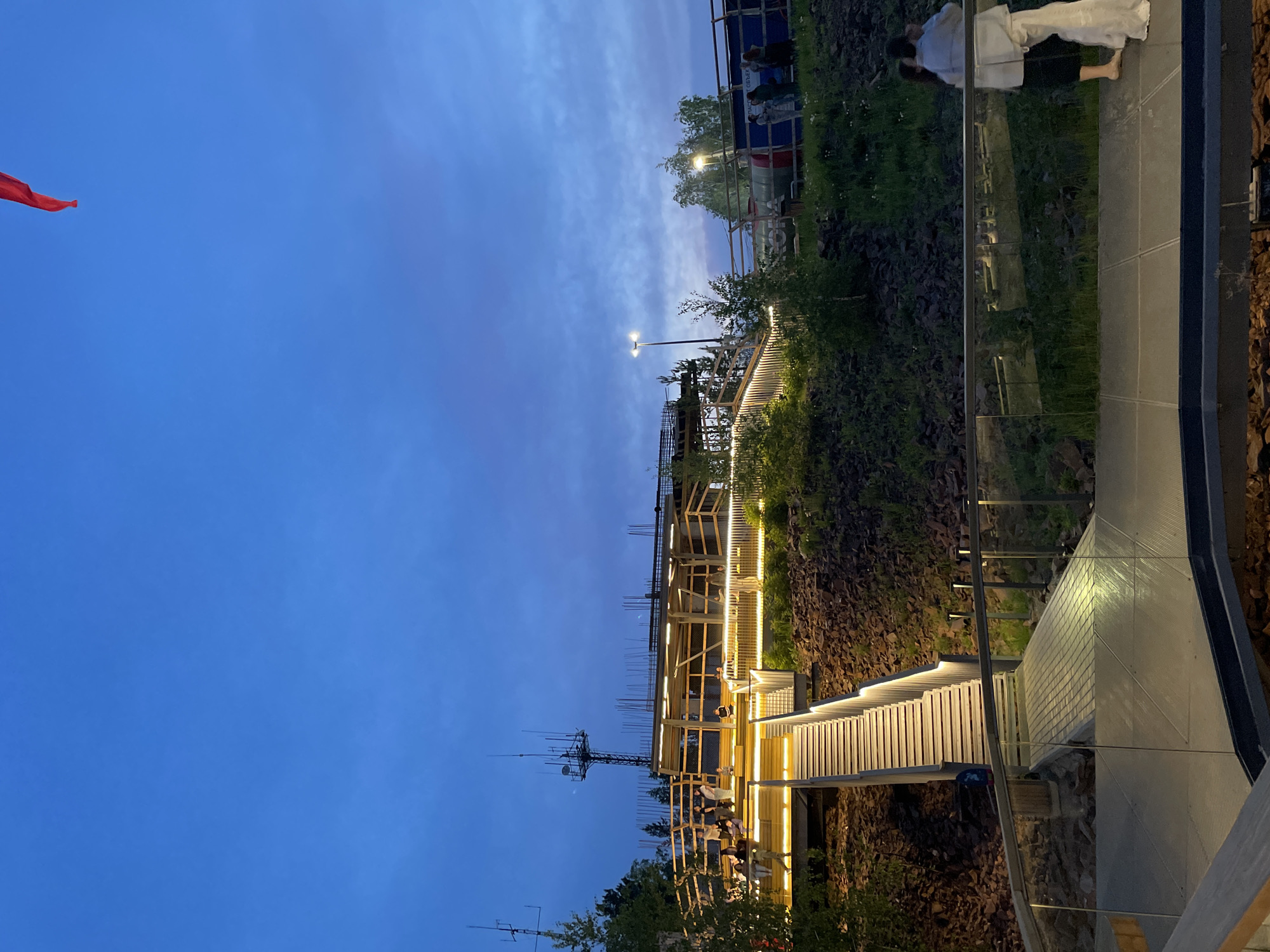 The image shows an outdoor amphitheater or viewing platform in Krasnoyarsk, Russia, at dusk. The structure is made of wood and metal, with illuminated stairs and railings. Several people are scattered throughout the area, some sitting on the steps and others standing and conversing. In the background, there is a construction site with scaffolding and a tall antenna tower. A red flag is visible in the top right corner of the image. The sky is a deep blue, indicating the transition from day to night, and the surrounding area is covered in greenery and rocks.