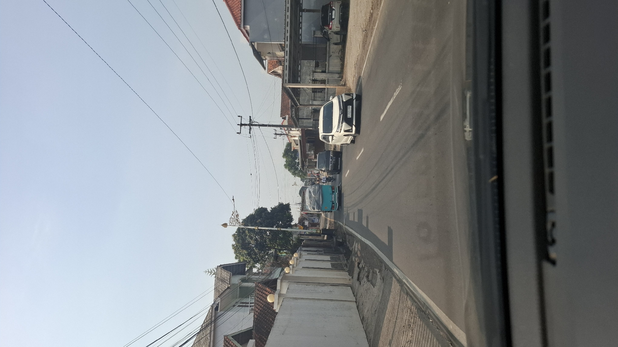 The image depicts a daytime scene on a paved road in Cilaku, Indonesia, likely taken from the perspective of a vehicle's dashboard. A white car is in the foreground, driving on the right side of the road. To the left of the road is a white wall with a tiled roof and a row of trees behind it. Further down the road, a turquoise truck with a load of grey material is visible, followed by a dark-colored car. The sky is clear and blue, with power lines crisscrossing overhead. Buildings line the right side of the road, featuring tiled roofs and covered carports. The road appears to be a two-lane highway with white lane markings.