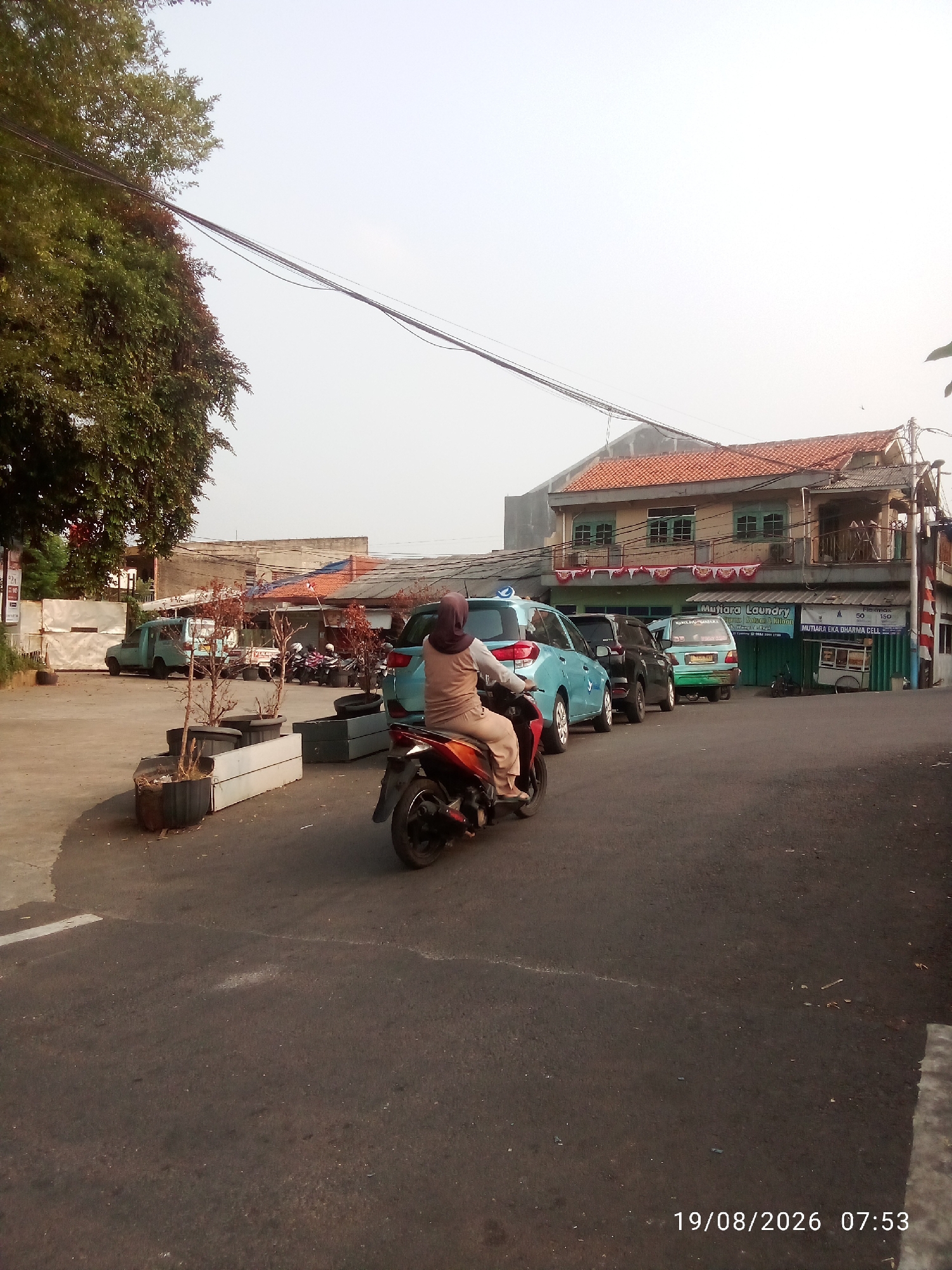 The image depicts a street scene in East Jakarta, Indonesia. In the foreground, a person on a motorcycle wearing a beige hijab and clothing is riding away from the camera. The motorcycle is orange and black. Behind the motorcycle, a line of vehicles is visible, including a blue car, a black SUV, and a green van. In the background, there are buildings with orange tiled roofs and signs, one of which reads "Mutiara Laundry". A large tree with green foliage is on the left side of the image, and power lines are visible in the sky. The ground is paved with asphalt, and there are some potted plants. The timestamp "19/08/2026 07:53" is displayed in the bottom right corner. The overall impression is a typical urban street with moderate traffic and commercial establishments.