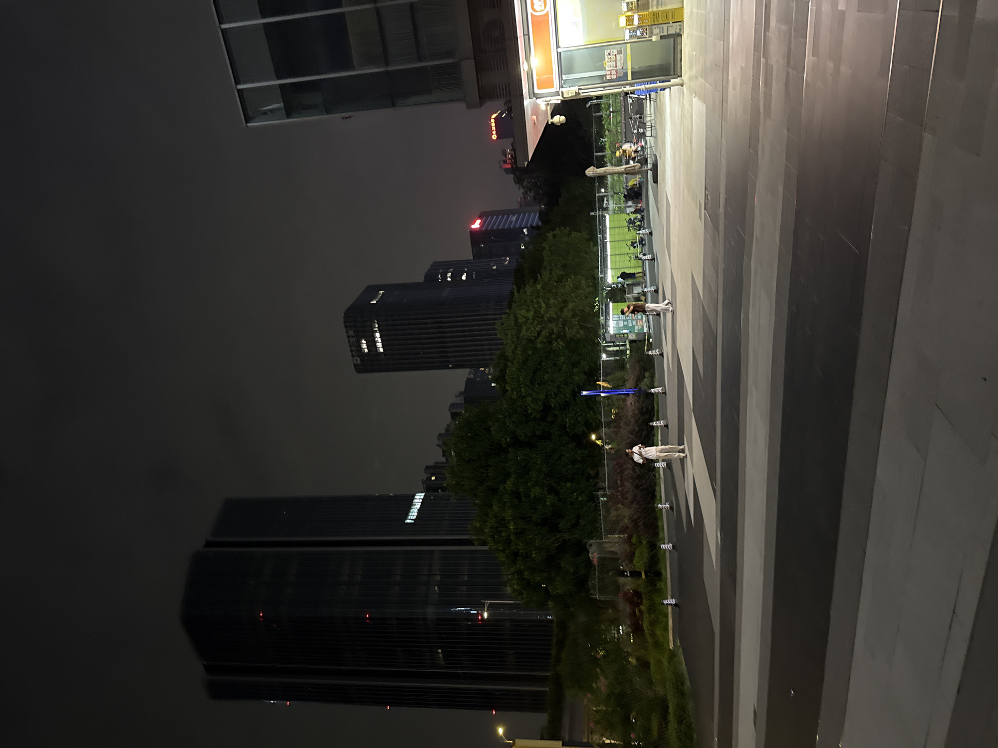 The image captures a nighttime scene in a modern urban environment, likely a plaza or walkway in Huangpu District, China. Dominating the background are several tall, dark skyscrapers with illuminated windows. In the foreground, a pedestrian walkway is marked by contrasting light and dark paving stones, creating a geometric pattern.
Several individuals are visible in the middle ground. One person, dressed in light-colored clothing, stands alone in the walkway, looking towards the camera. Further back, near a fenced-off green area, a small group of people are gathered, some seated at tables, and others standing. Bicycles are parked nearby, suggesting a public transportation or recreational area.
To the right of the scene, a section of a modern building with large glass windows is visible. Adjacent to it is a small kiosk-like structure with yellow signage and a display of products, possibly a convenience store or vendor stall. The overall atmosphere is one of urban night life, with the city lights and structures illuminated against the dark sky.