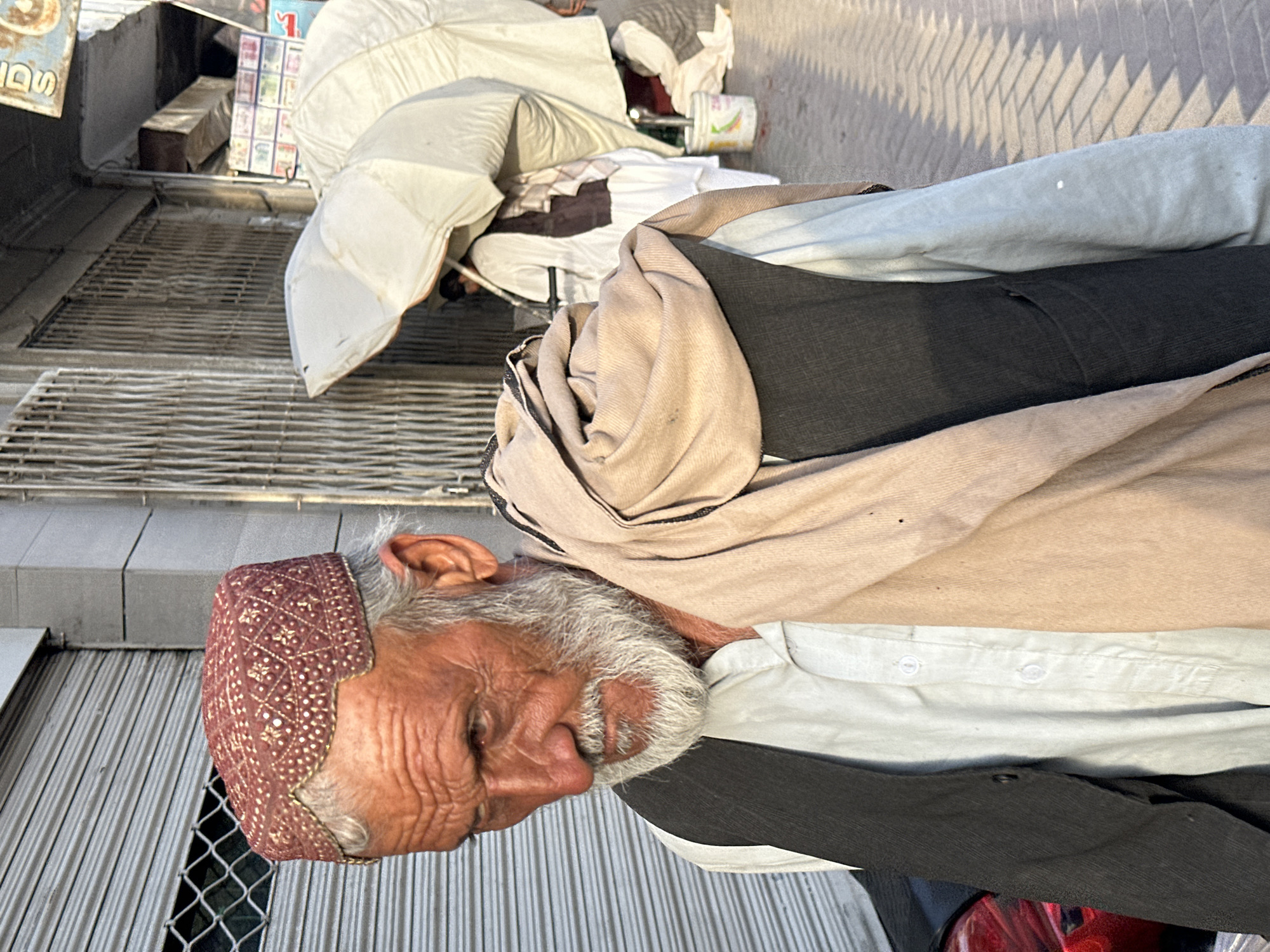The image depicts an elderly man wearing a red and gold patterned skullcap. He has gray hair and beard, and his face is lined with age, giving him a thoughtful expression. He is dressed in traditional clothing, including a light-colored shirt and a dark vest. A tan-colored shawl is draped around his neck.
The man appears to be sitting or resting, possibly in an outdoor market or street setting. Behind him, there are indications of stalls or shops with metal shutters and a wire mesh fence. In the background, there are some scattered objects, including white fabric, possibly part of a stall or awning, and some boxes or crates. The overall scene suggests a casual daytime environment.
The lighting indicates it is daytime, with shadows cast on the ground, possibly indicating mid-morning or afternoon. The texture of the ground suggests it might be paved or a gritty surface. The man's posture and facial expression do not convey any strong emotions; he appears to be relaxed or perhaps contemplative.
There is some text visible in the upper left corner of the image on a sign, but it is partially obscured and difficult to read clearly. It appears to be in a script common in Pakistan. The broader context of Quetta City Tehsil, Pakistan, places the scene within a specific cultural and geographical setting.