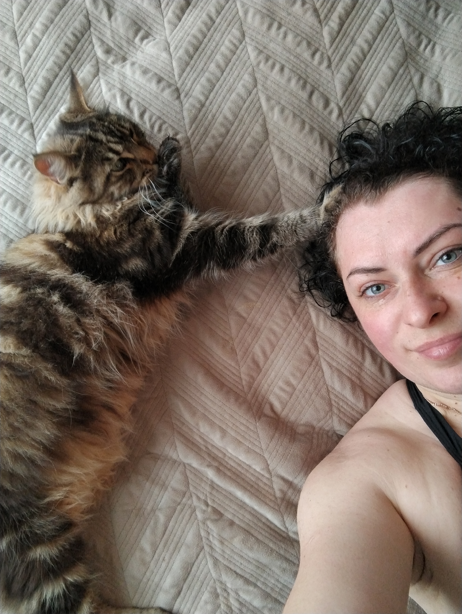 A woman with curly black hair and light blue eyes lies on a quilted beige bedspread next to a tabby cat with long fur. The cat's paw is raised near the woman's face. The woman is wearing a black top, and a gold necklace is visible. No information indicates this photo was taken in the city of Grishanki, Russia.