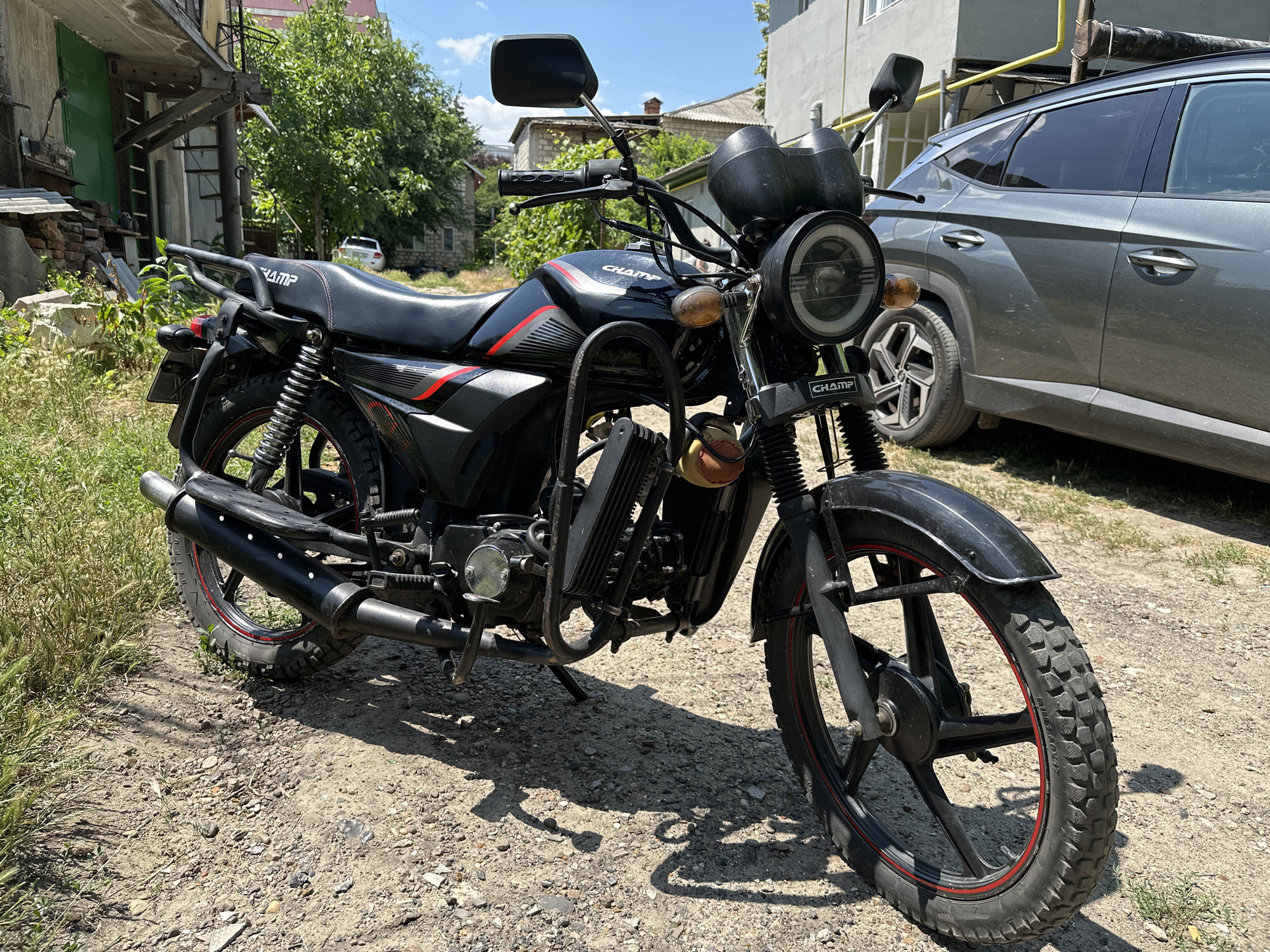 A black motorcycle with red accents is parked on a gravelly surface outdoors, near a building and a grey SUV. The motorcycle features a round headlight with a halo ring, dual mirrors, and "CHAMP" branding on the seat and front. The background shows a building with a light-colored facade and windows, trees, and a clear blue sky. The ground is covered in loose gravel and some dry grass. The overall scene suggests an urban or suburban environment in Comrat, Moldova, likely during daylight. There are no people visible in the image.