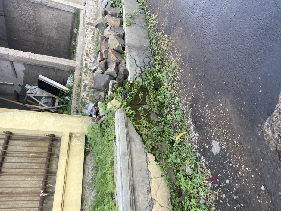 The image shows a section of a concrete sidewalk in Bandung, Indonesia, at the coordinates -6.938 latitude and 107.558 longitude. The sidewalk is cracked and uneven, with green plants growing up through the cracks. There are several rocks piled up next to the sidewalk. In the background, a yellow building with a barred window is visible. The sidewalk is adjacent to an asphalt road. A piece of yellow debris is visible in the lower left corner of the image. The concrete shows signs of weathering and erosion.